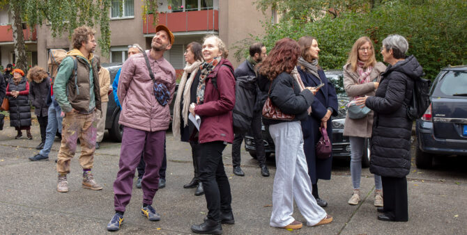 Frauen und Männer verschiedenen Alters stehen in einer Wohnsiedlung und unterhalten sich, einige von Ihnen schauen nach oben zu einer Häuserfassade. Foto: Ludger Paffrath