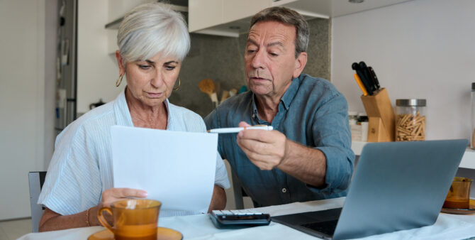 Ein älteres Paar sitzt am Küchentisch, bespricht wichtige Dokumente und Finanzen, mit Laptop und Taschenrener