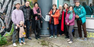 Eine Gruppe Menschen posiert an einer großen, runden Regentonne und lächelt in die Kamera. Zwei Personen halten einen Gießkanne. Foto: Ralph Maak