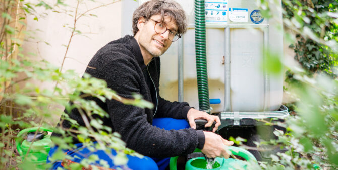Ein Mann mit Brille hockt in vor einer Regentonne und zapft Wasser in eine Gießkanne. Foto: Ralph Maak