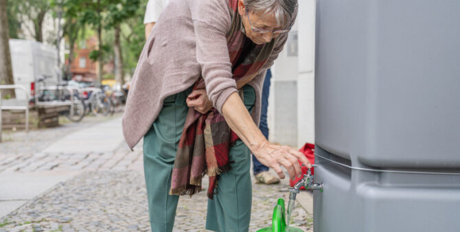 Eine Frau bedient den Hahn einer Regentonne, die vor einer Häuserfassade steht, und füllt Wasser in eine Gießkanne. Foto: Felix Seyfert