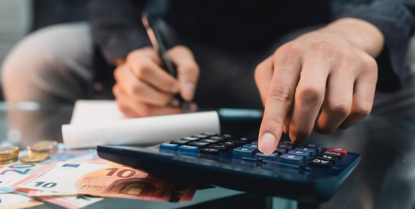 Detailaufnahme von Händen, die auf einem Taschenrechner tippen, umgeben von Euro-Geld und Notizen