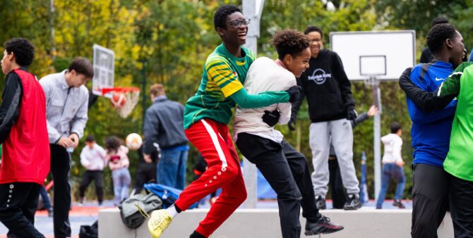 Szene auf einem Bolzplatz voller Kinder. Im Fokus: Zwei Jungs, die sich springend umarmen und lachen. Foto: City-Press GmbH