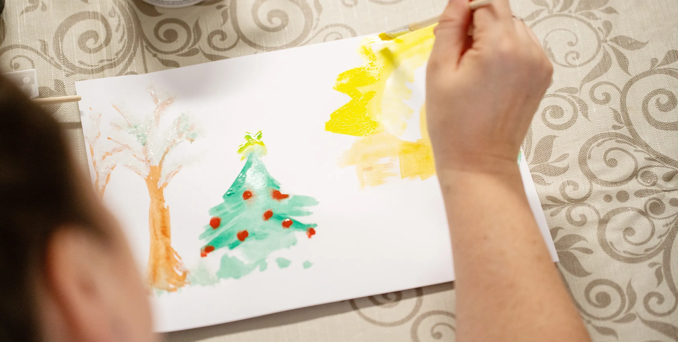Blick über die Schulter: Eine Person malt mit Wasserfarbe und Pinsel ein weihnachtliches Bild, auf dem u.a. ein Christbaum zu sehen ist. Foto: Ralph Maak