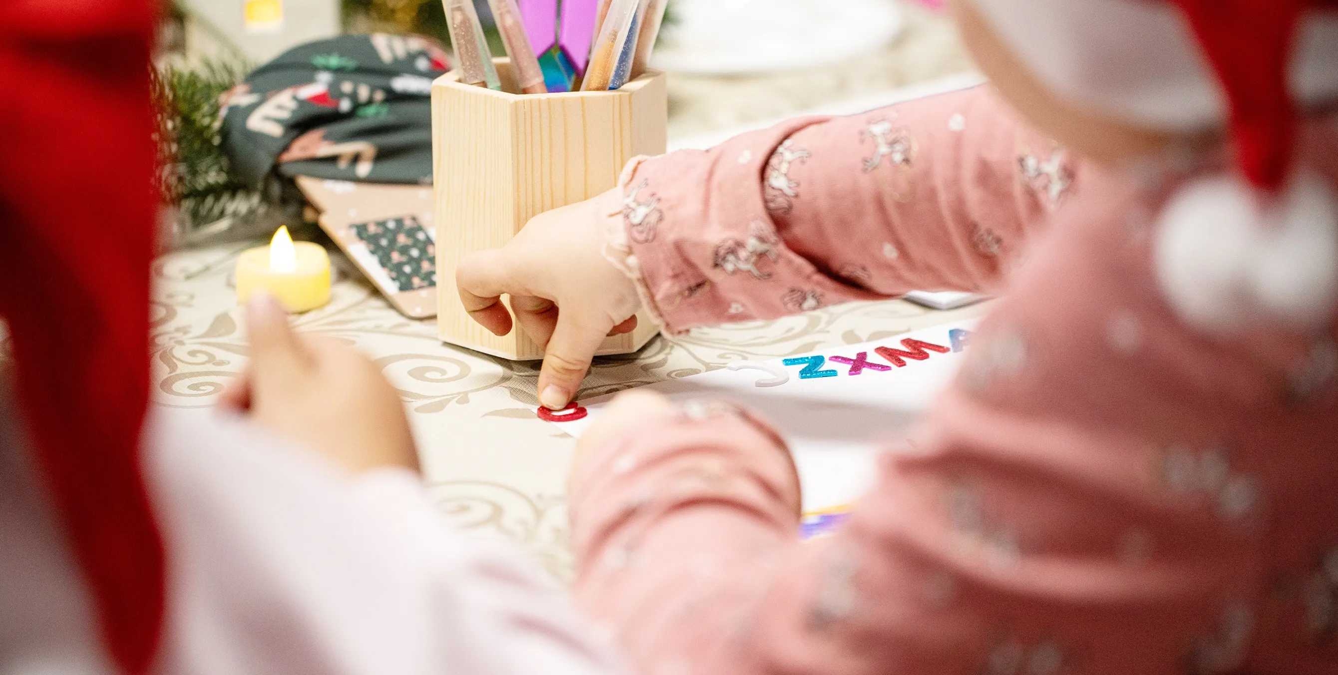 Großaufnahme: Eine Kinderhand drückt einen Klebebuchstaben auf ein Blatt Papier. Im Hintergrund sind Filzstifte und Teelichter zu sehen. Foto: Ralph Maak