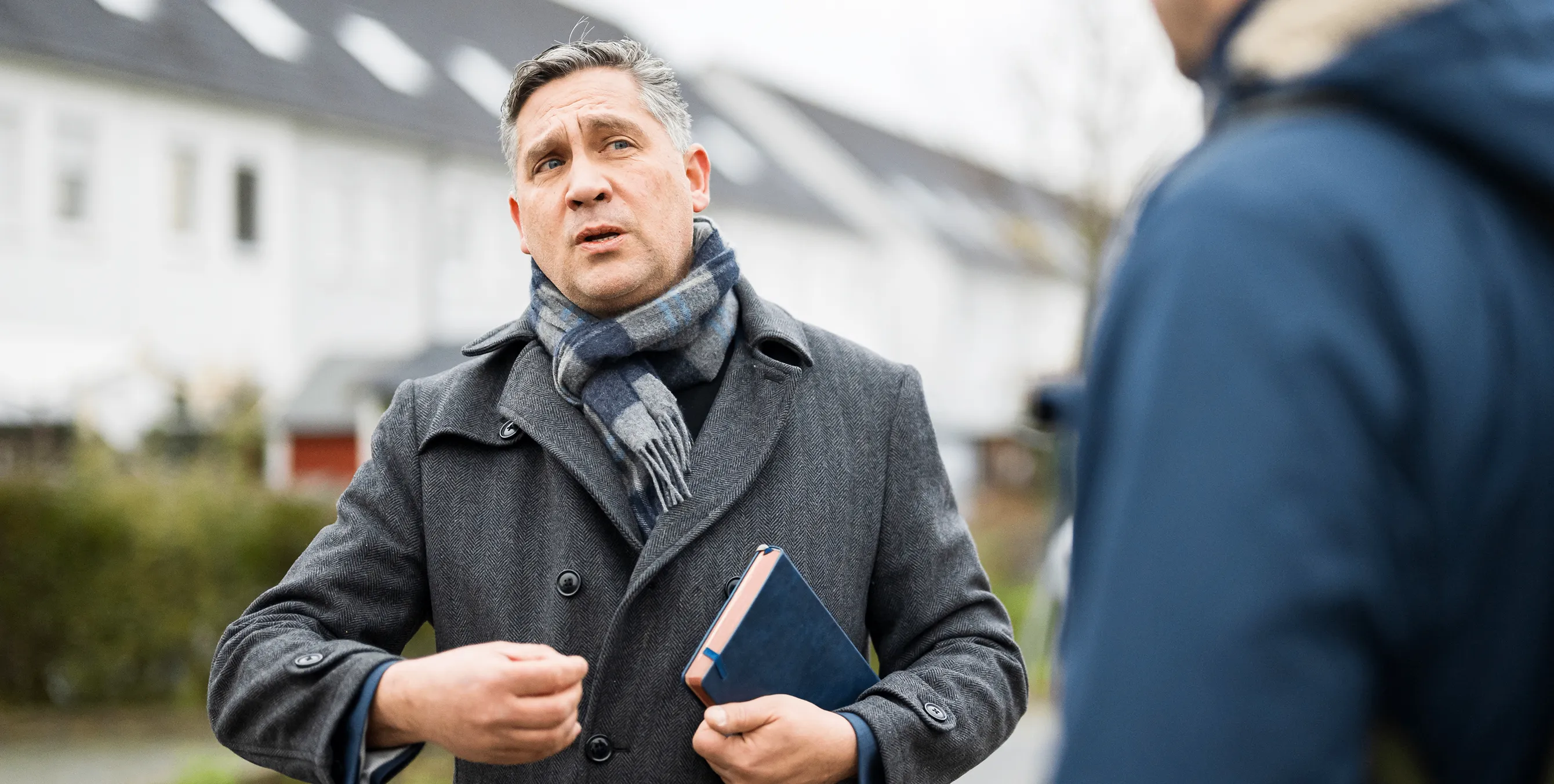 Ein Mann in grauem Mantel und Schal steht in einem Wohnquartier und spricht zu einer anderen Person. Foto: City-Press GmbH