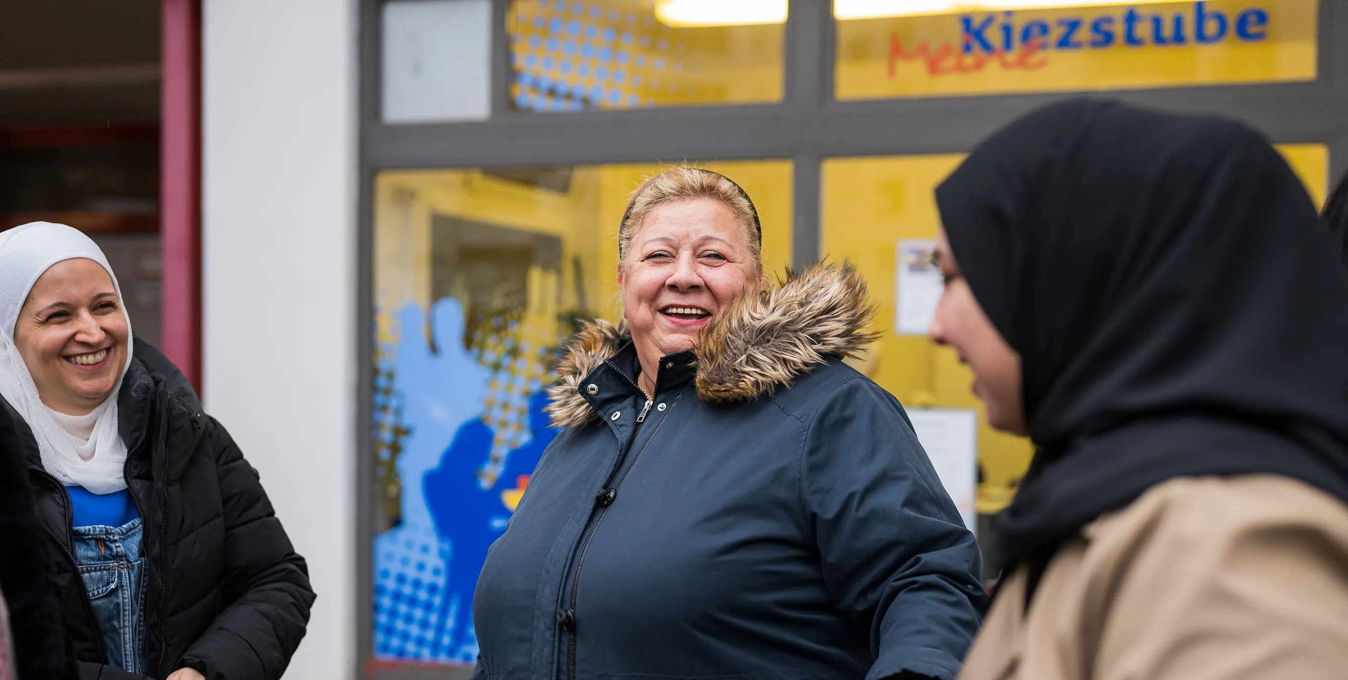 Eine ältere Dame steht in Winterjacke im Freien und unterhält sich lächelnd mit zwei jüngeren Frauen, die Kopftücher tragen. Im Hintergrund ist ein Ladenfenster mit der Aufschrift "Meine Kiezstube" zu sehen. Foto: City-Press GmbH