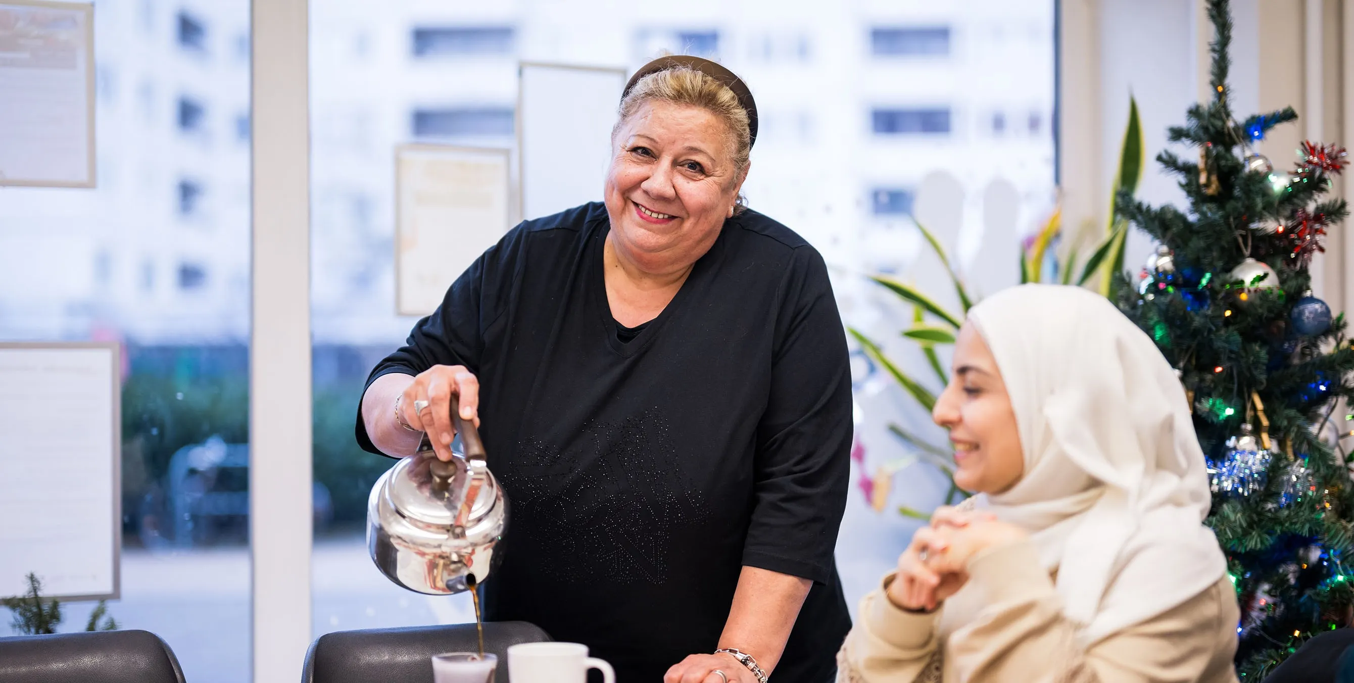 Eine ältere Dame steht in einem weihnachtlich geschmückten Raum an einem Tisch und schenkt einer jüngeren, sitzenden Frau mit Kopftuch Tee ein. Foto: City-Press GmbH