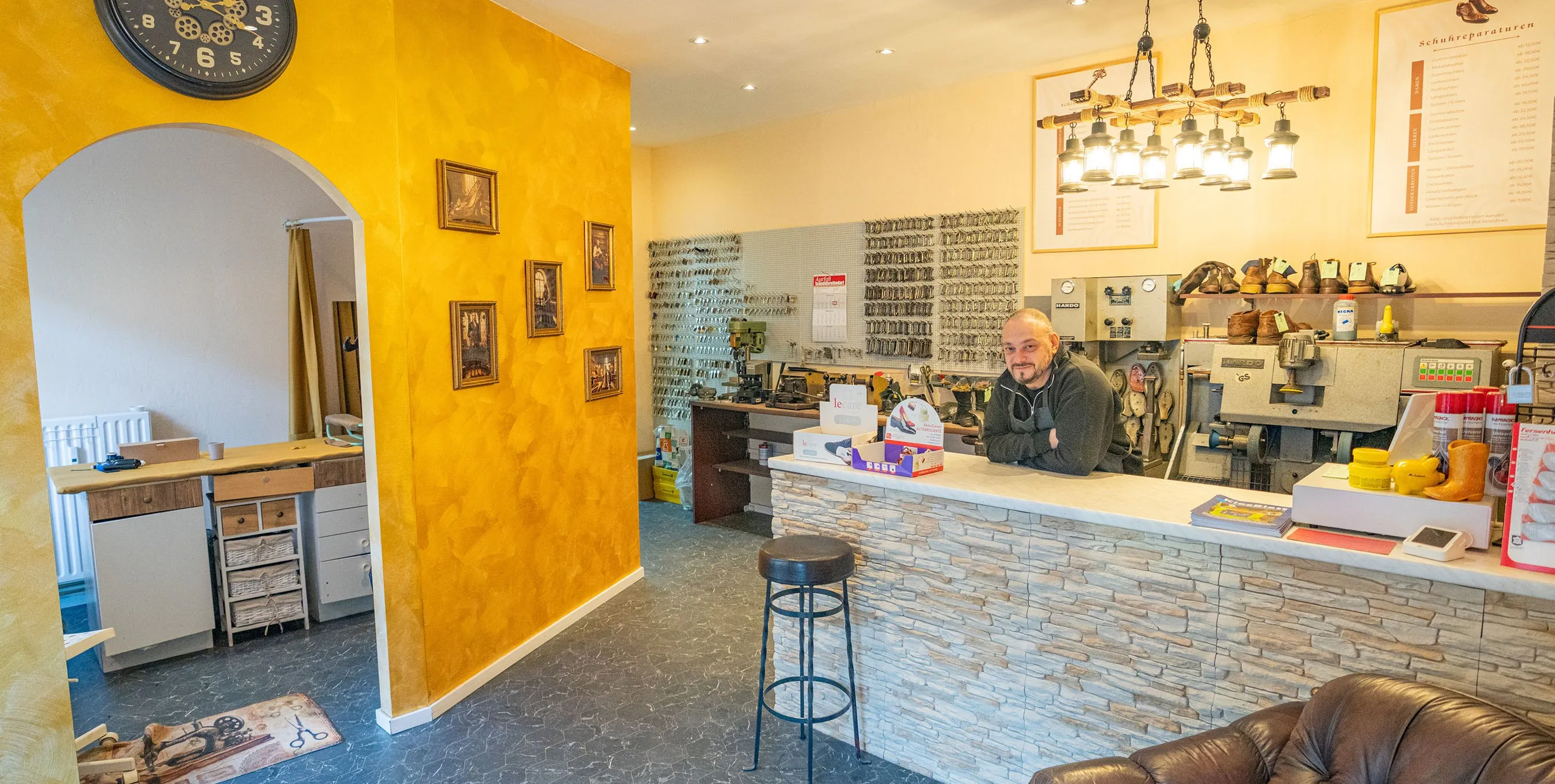 Ein Schuhmacher lehnt in seinem Ladengeschäft über dem Tresen. Im Hintergrund sind Schuhe, Maschinen und Schlüsselrohlinge zu sehen. Foto: Felix Seyfert