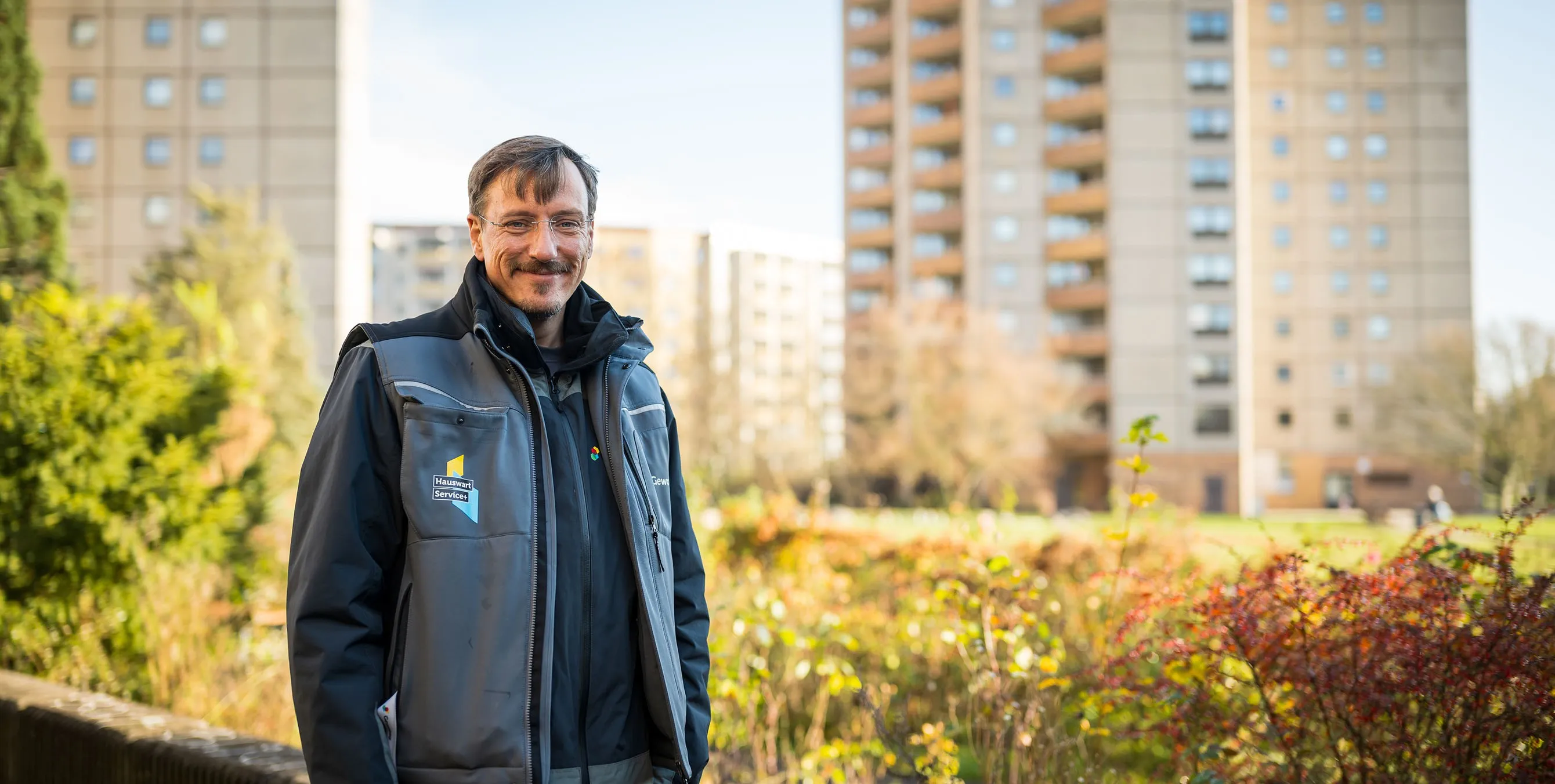 Ein Hauswart steht im Freien vor einer Grünfläche und lächelt in die Kamera. Im Hintergrund sind Hochhäuser zu sehen. Foto: City-Press GmbH