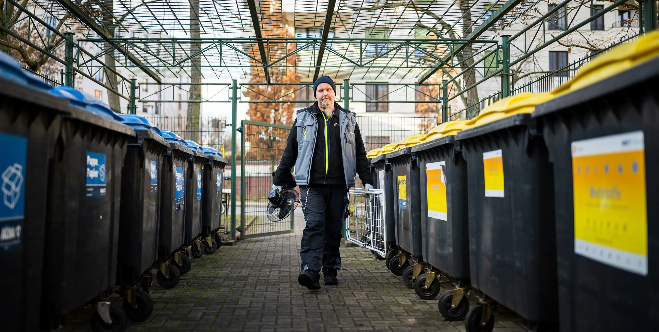 Ein Hauswart trägt zwei kleine Sperrmüllgegenstände in einen eingezäunten Müllplatz mit zahlreichen Mülltonnen. Foto: City-Press GmbH