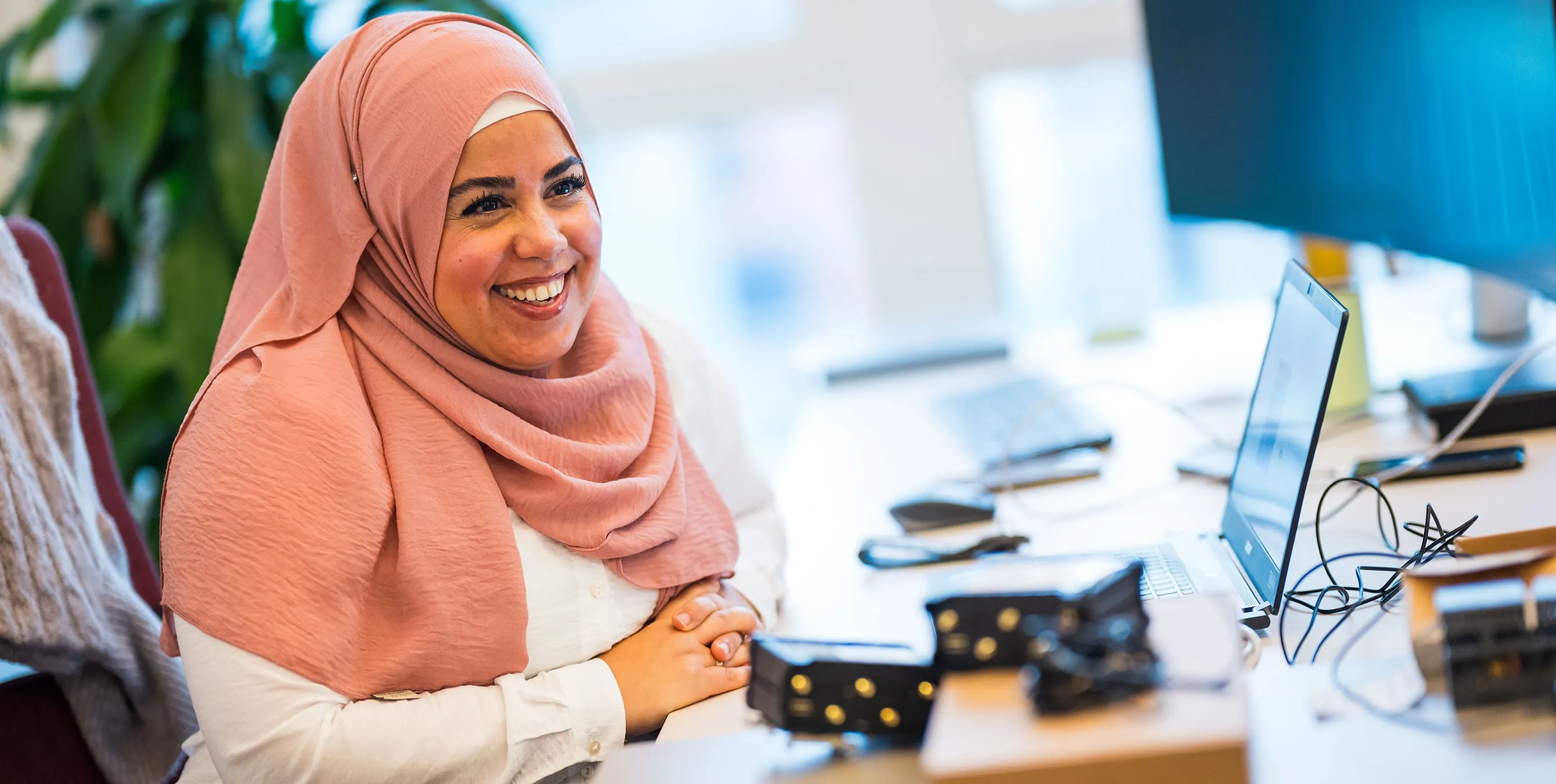 Eine lächelnde Frau mit Kopftuch sitzt an einem Arbeitsplatz mit Laptop und großem Bildschirm. Foto: City-Press GmbH