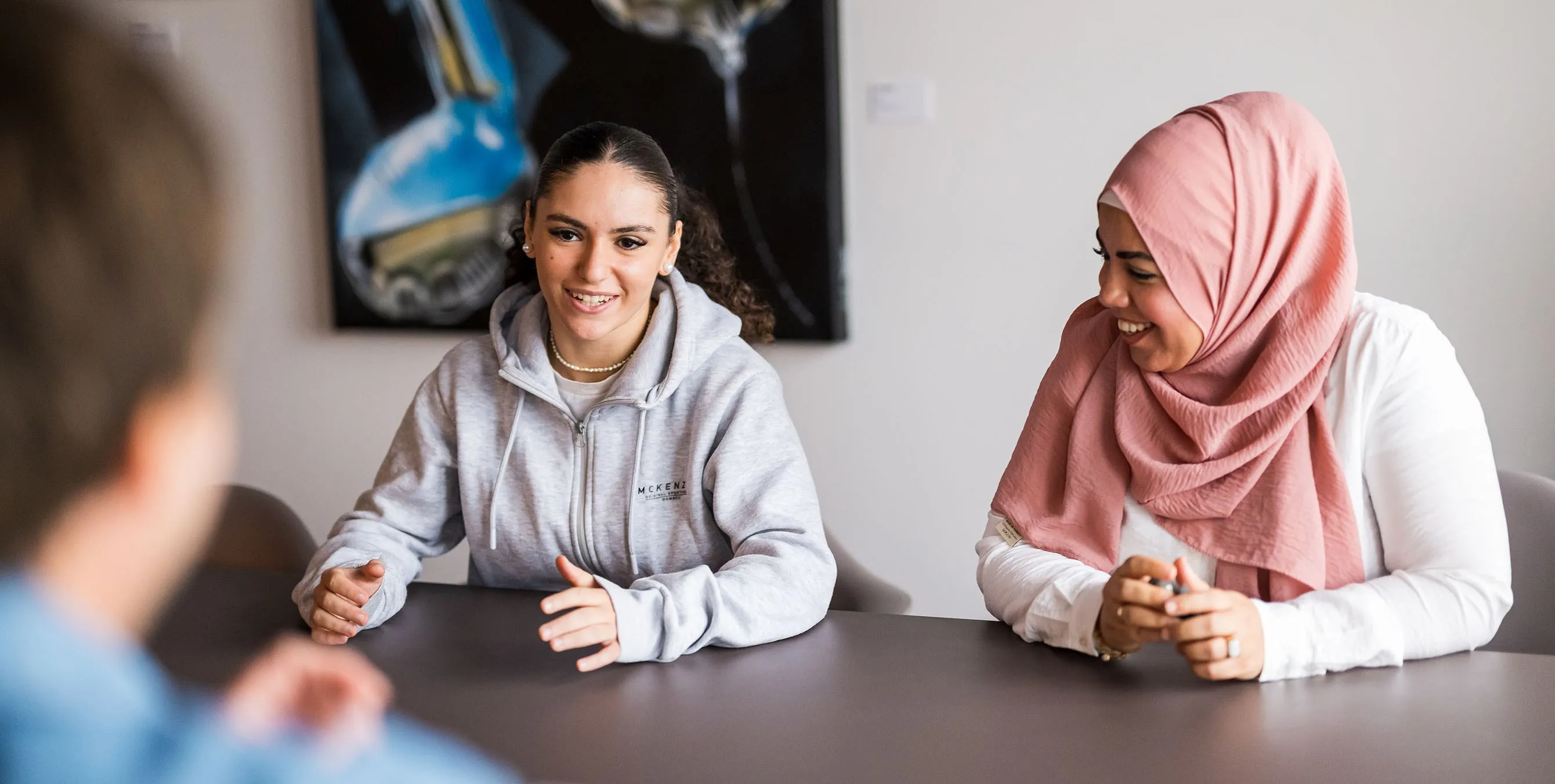 Zwei Frauen sitzen an einem Tisch, lächeln und sprechen mit einer gegenübersitzenden Person. Foto: City-Press GmbH