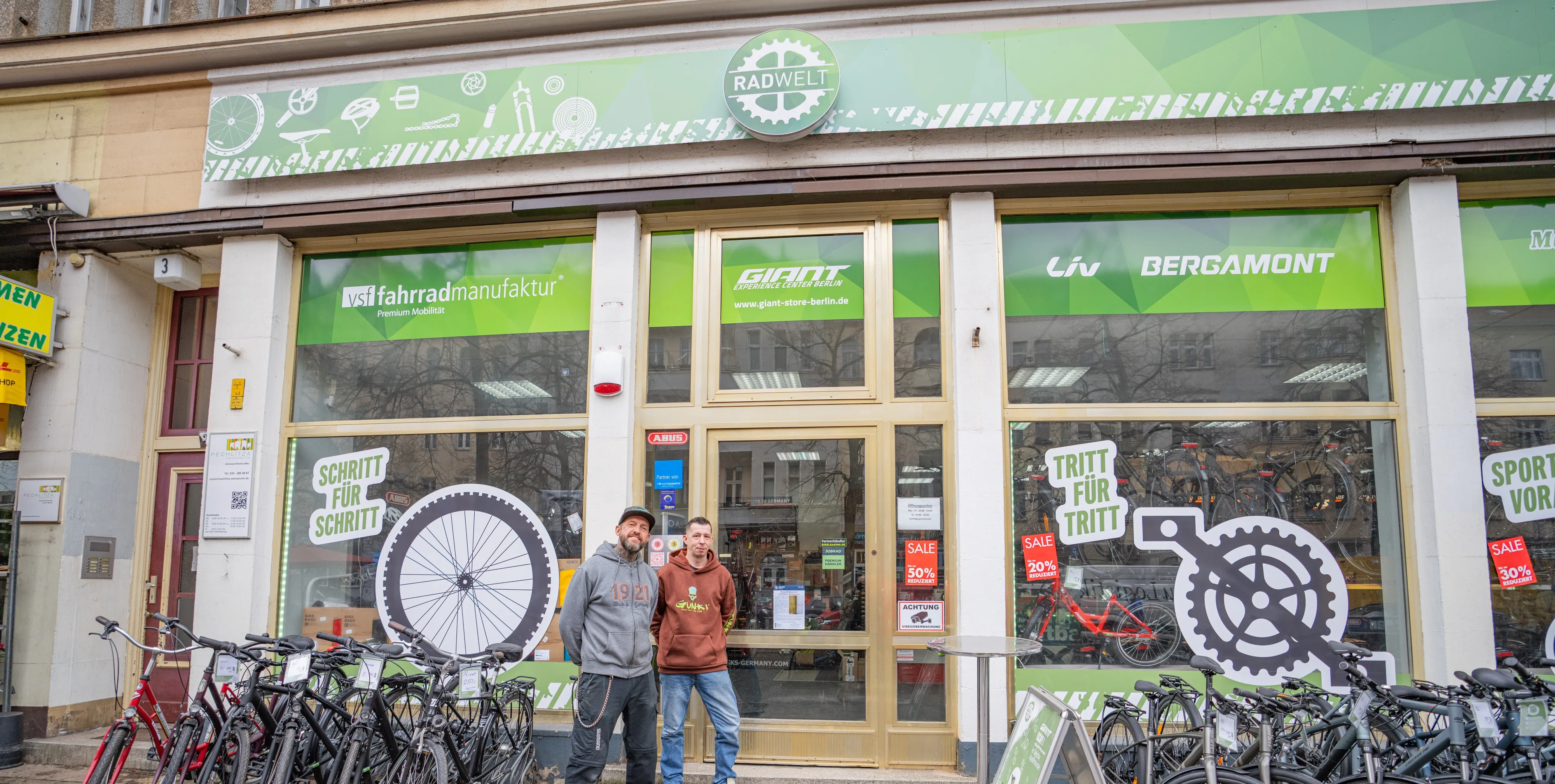 Zwei Männer stehen vor einem Fahrradladen in Berlin-Pankow zwischen einigen Fahrrädern. Der Laden heißt Radwelt.