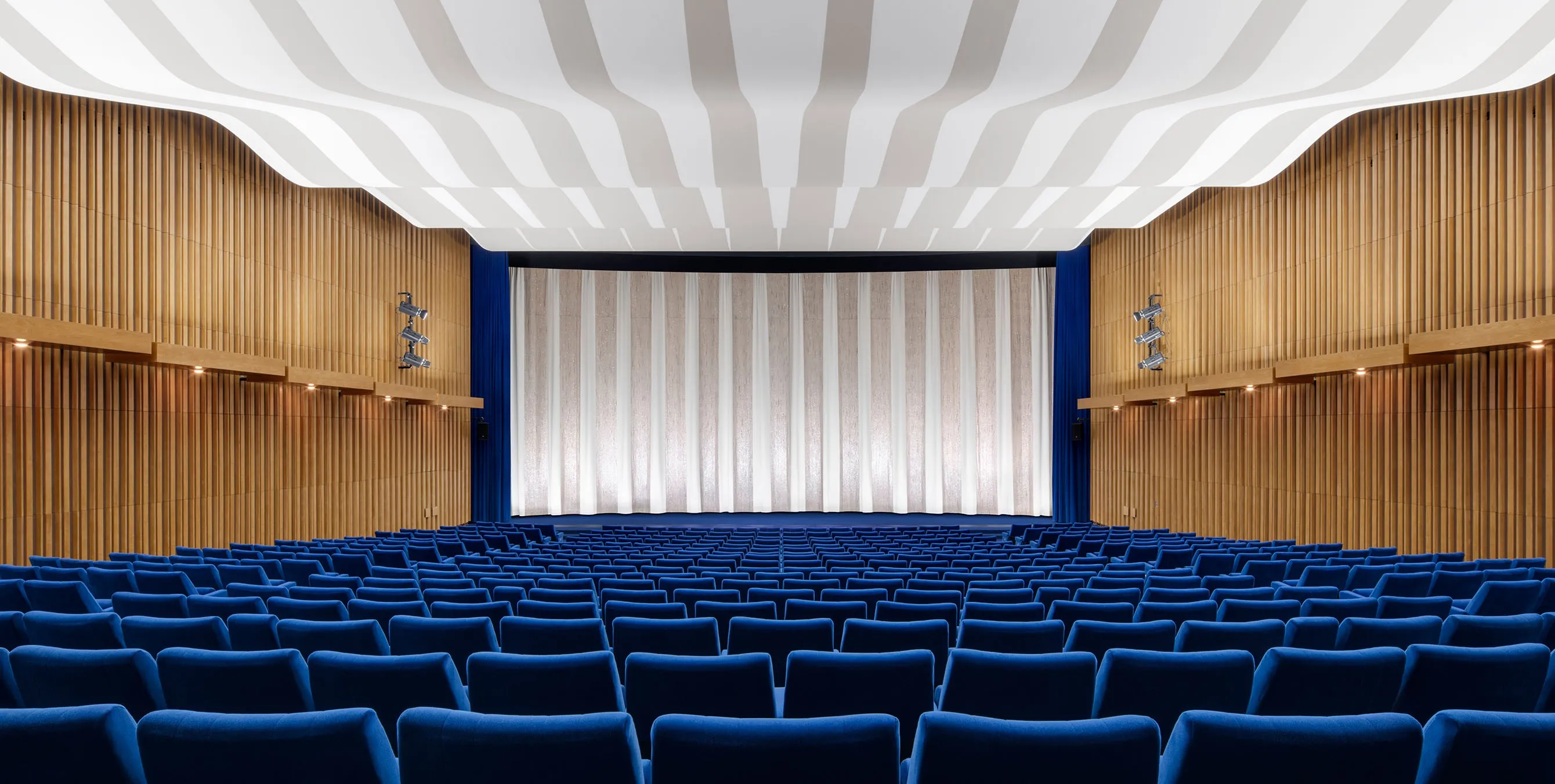 Ansicht von hinten: Ein großer, menschenleerer Kinosaal mit blauen Sitzen, holzvertäfelten Wänden und geschwungener Deckenkonstruktion. Foto: Yorck Kinogruppe/Daniel Horn