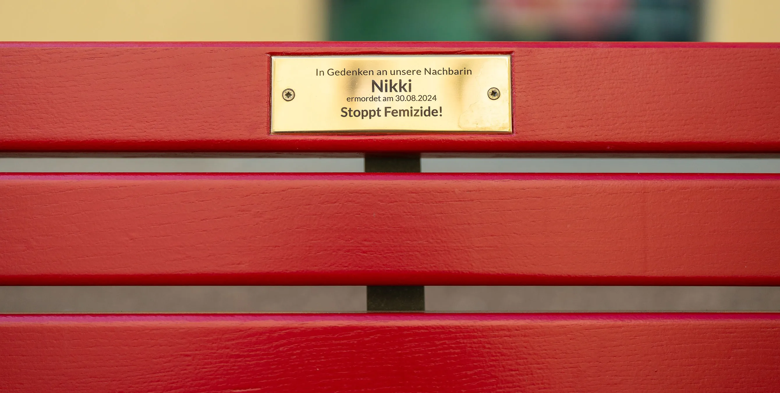 Großaufnahme: Eine rote Sitzbank mit einer goldenen Plakette an der Sitzlehne. Aufschrift: In Gedenken an unsere Nachbarin Nikki, ermordet am 30.08.2024. Stoppt Femizide! Foto: Ralph Maak
