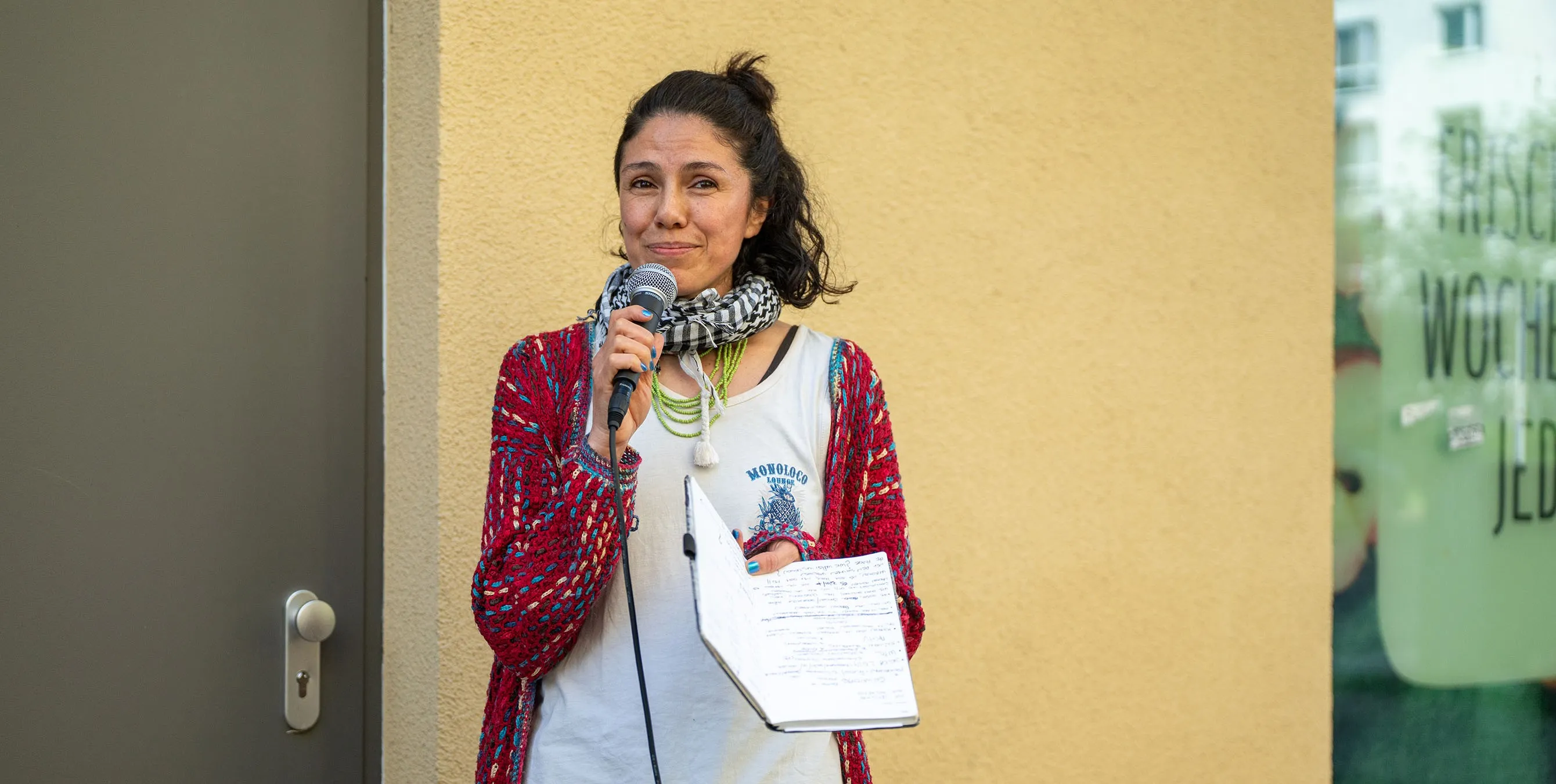 Eine Frau mit dunklen Haaren steht vor einer gelben Wand. IN der einen Hand hält sie ein Notizbuch, in der anderen ein Mikrofon, mit dem sie eine Rede hält. Foto: Ralph Maak