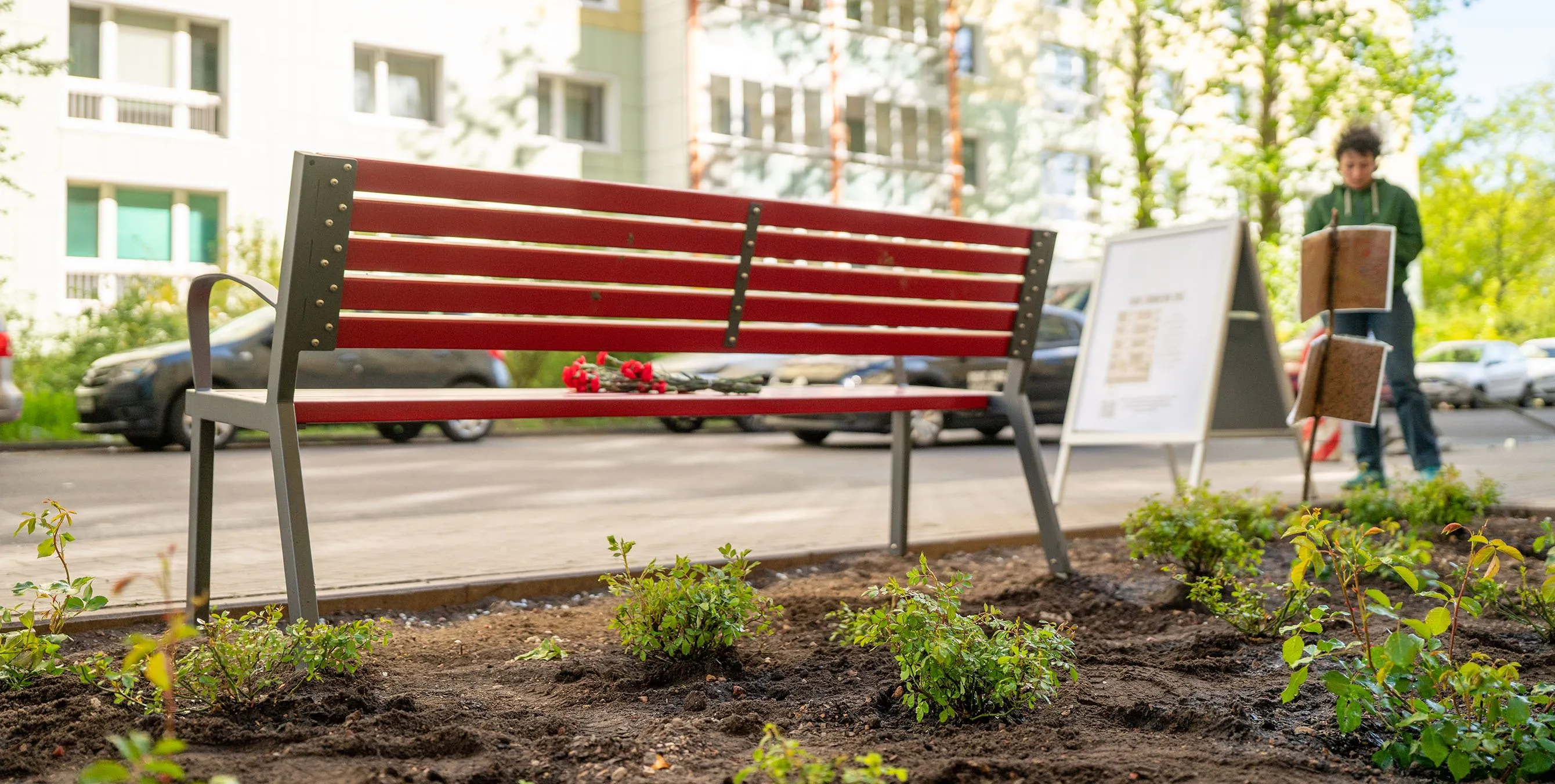 Aufnahme von hinten: Eine rote Sitzbank am vorderen Ende eines frisch bepflanzten Beets. Auf der Sitzfläche der Bank liegt ein Blumenstrauß. Foto: Ralph Maak