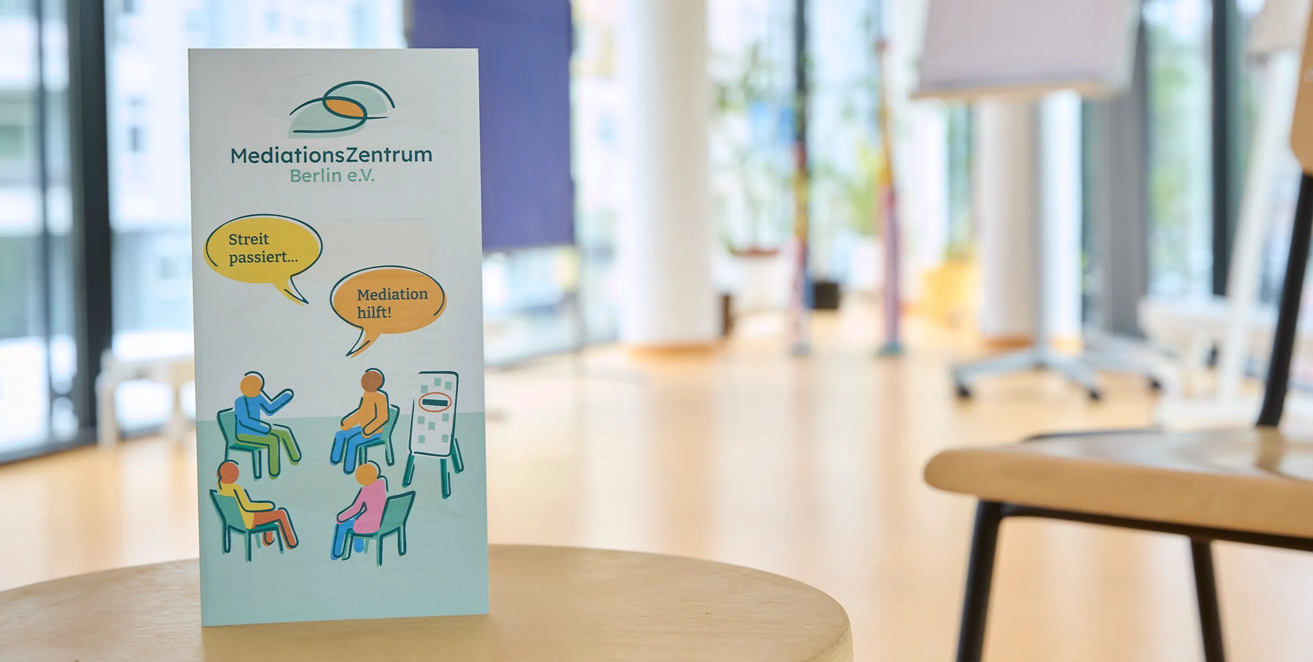 Großaufnahme: Ein Infoflyer steht in einem Besprechungsraum auf einem Tisch. Auf dem Flyer ist zu lesen: "MediationsZentrum Berlin e. V. Streit passiert, Mediation hilft!" Foto: Britta Leuermann