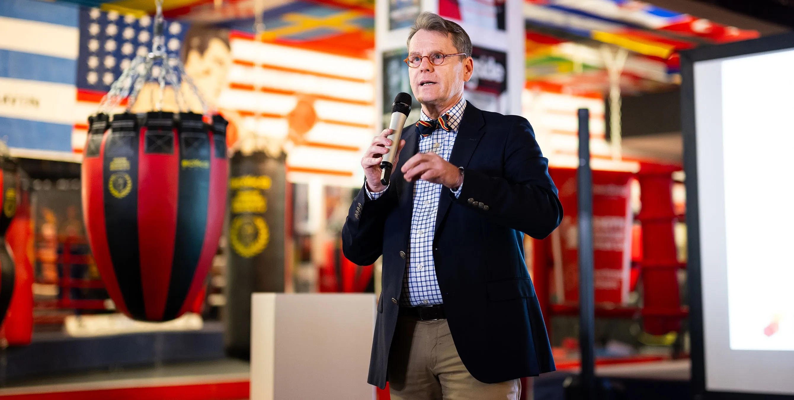 Ein Herr mit Brille und Fliege hält ein Mikrofon in der Hand und hält eine Rede in einem Boxgym. Foto: City-Press GmbH