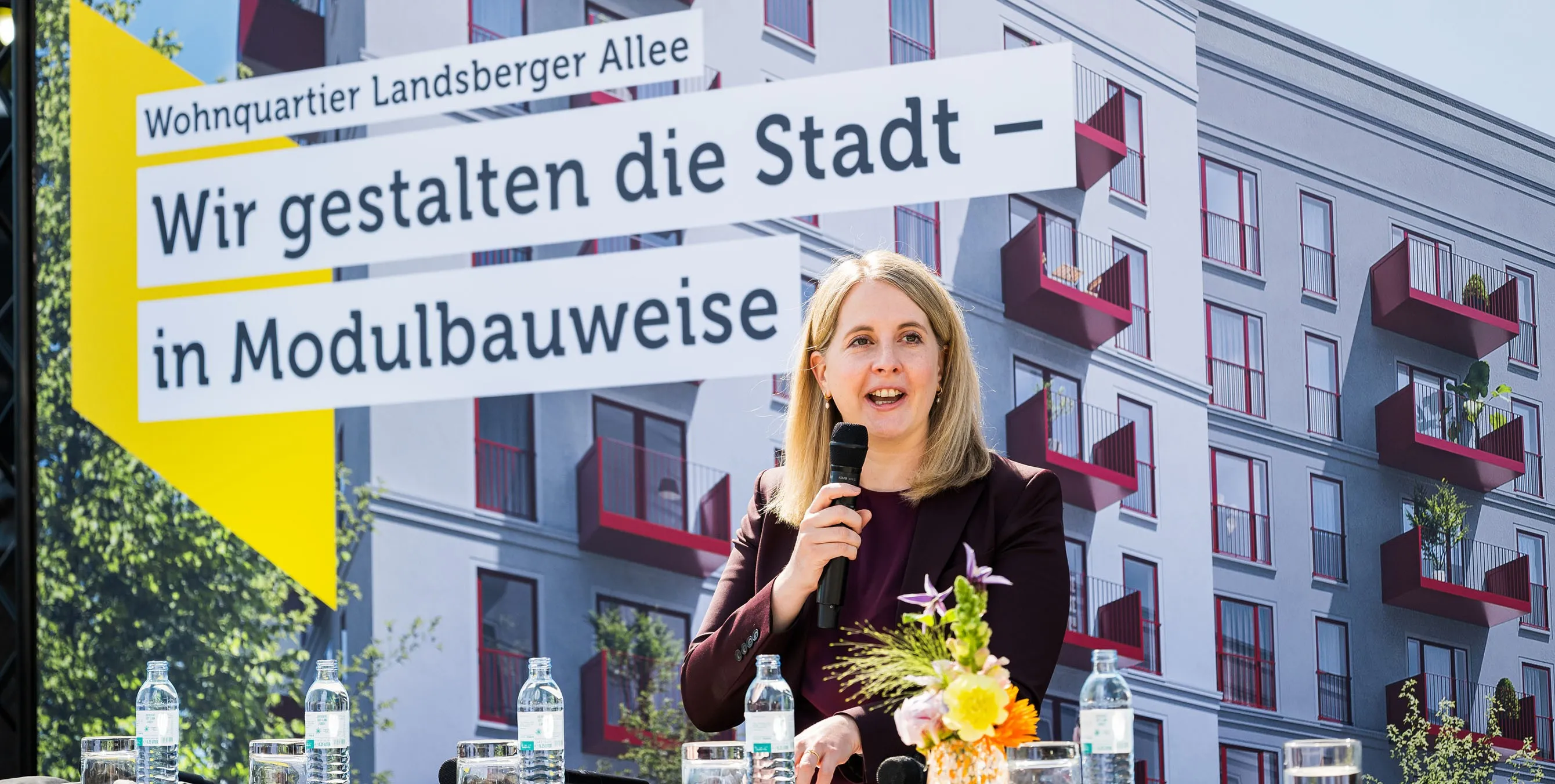 Ein Frau steht im Freien auf einer Bühne und spricht in ein Mikrofon. Im Hintergrund ist ein großes Bild eines Neubaus zu sehen, dazu der Schriftzug: Wir gestalten die Stadt - in Modulbauweise. Foto: City-Press GmbH