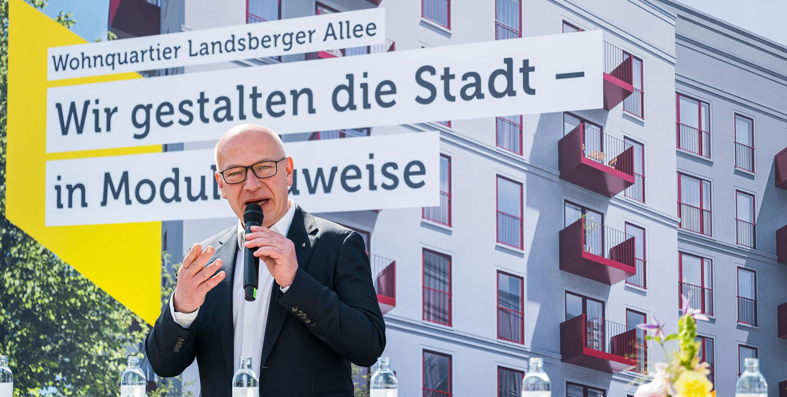 Ein Mann mit Brille steht im Freien auf einer Bühne und spricht in ein Mikrofon. Im Hintergrund ist ein großes Bild eines Neubaus zu sehen, dazu der Schriftzug: Wir gestalten die Stadt - in Modulbauweise. Foto: City-Press GmbH