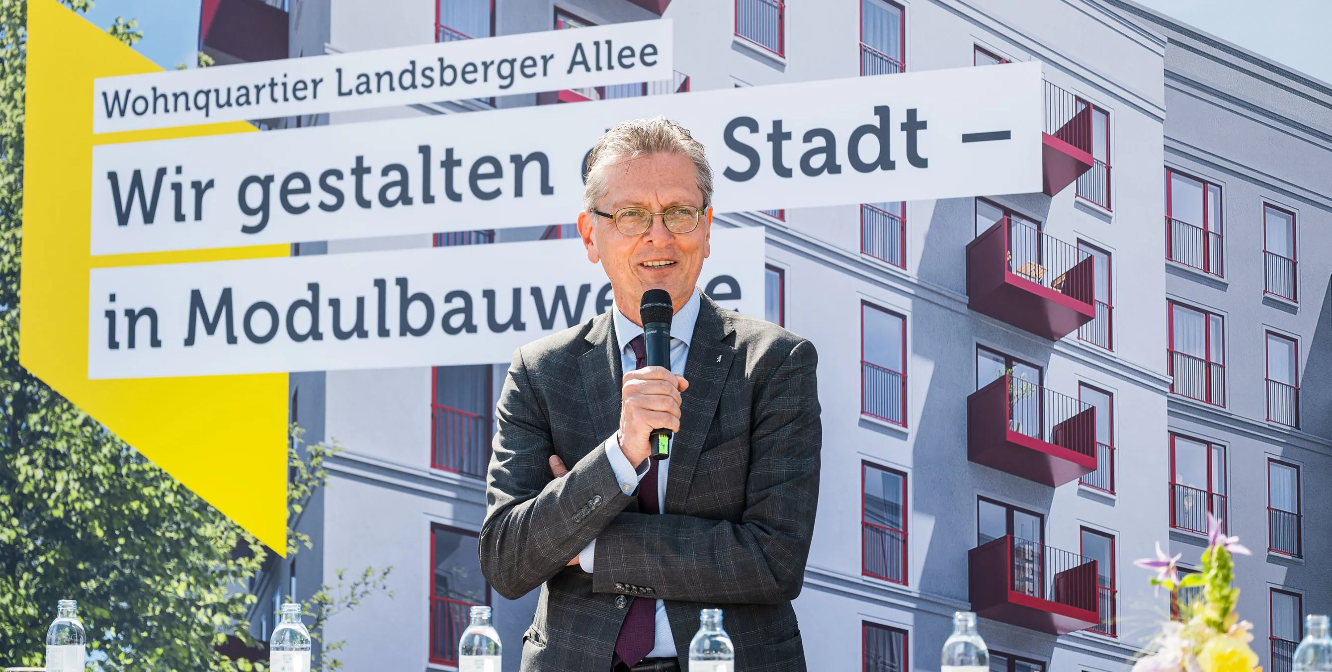 Ein Mann mit Brille steht im Freien auf einer Bühne und spricht in ein Mikrofon. Im Hintergrund ist ein großes Bild eines Neubaus zu sehen, dazu der Schriftzug: Wir gestalten die Stadt - in Modulbauweise. Foto: City-Press GmbH