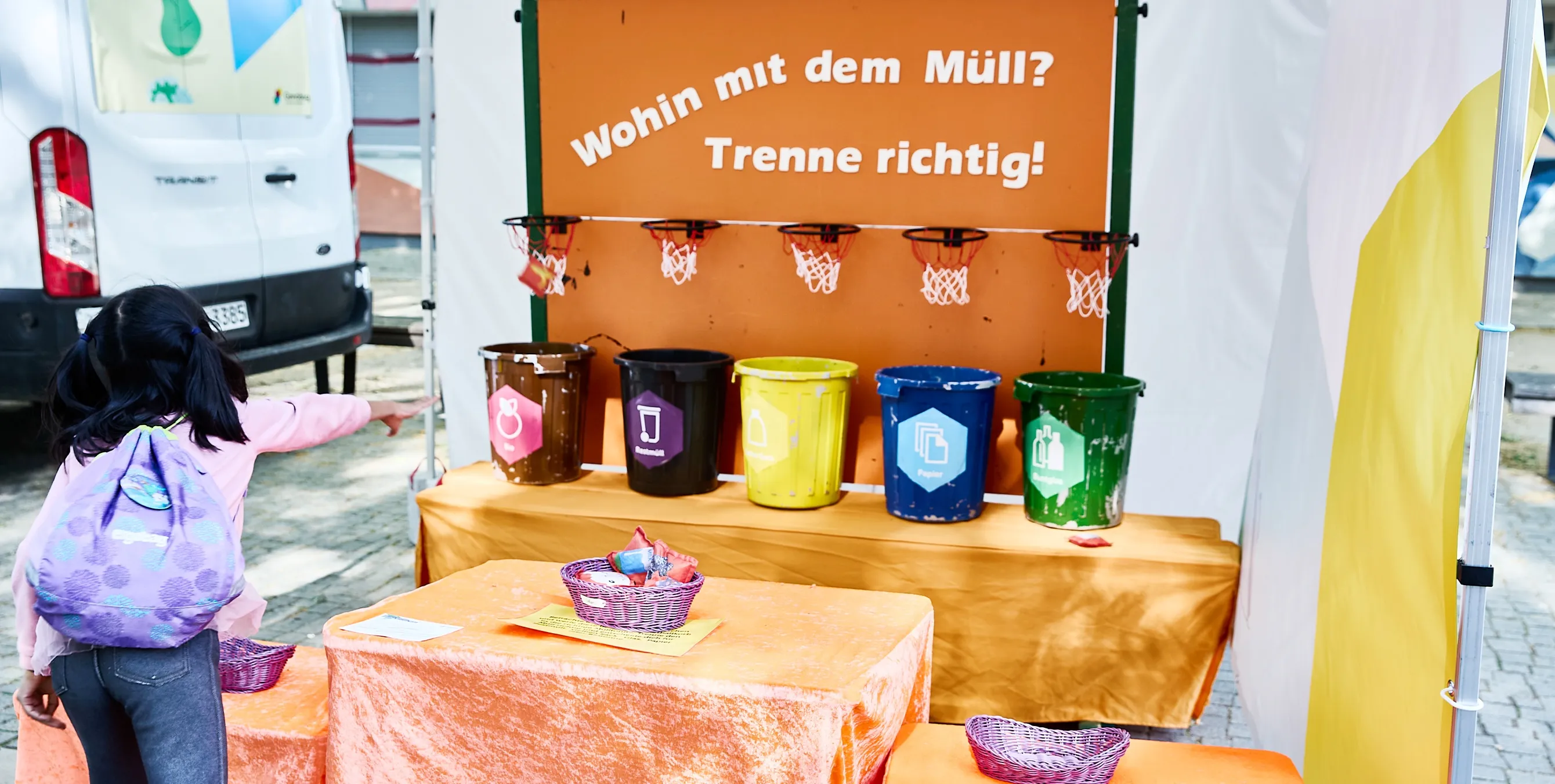 Ein Mädchen an einem Mitmachstand für Kinder, bei dem Gegenstände durch kleine Basketballkörbe in verschiedene Mülleimer geworfen werden müssen. An dem Stand prangt der Schriftzug: Wohin mit dem Müll? Trenne richtig! Foto: Britta Leuermann