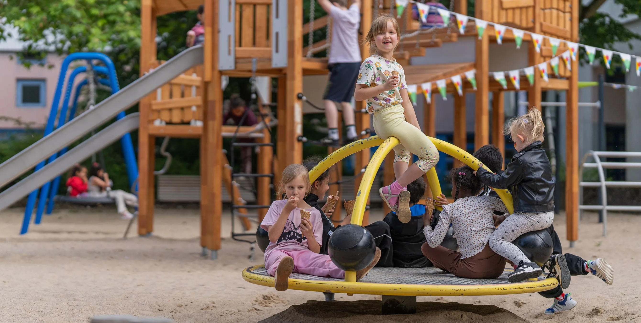 Szene auf einem Spielplatz im Freien: Mehrere Kinder sitzen auf einem Drehkarussell und essen dabei Eis. Foto: Felix Seyfert
