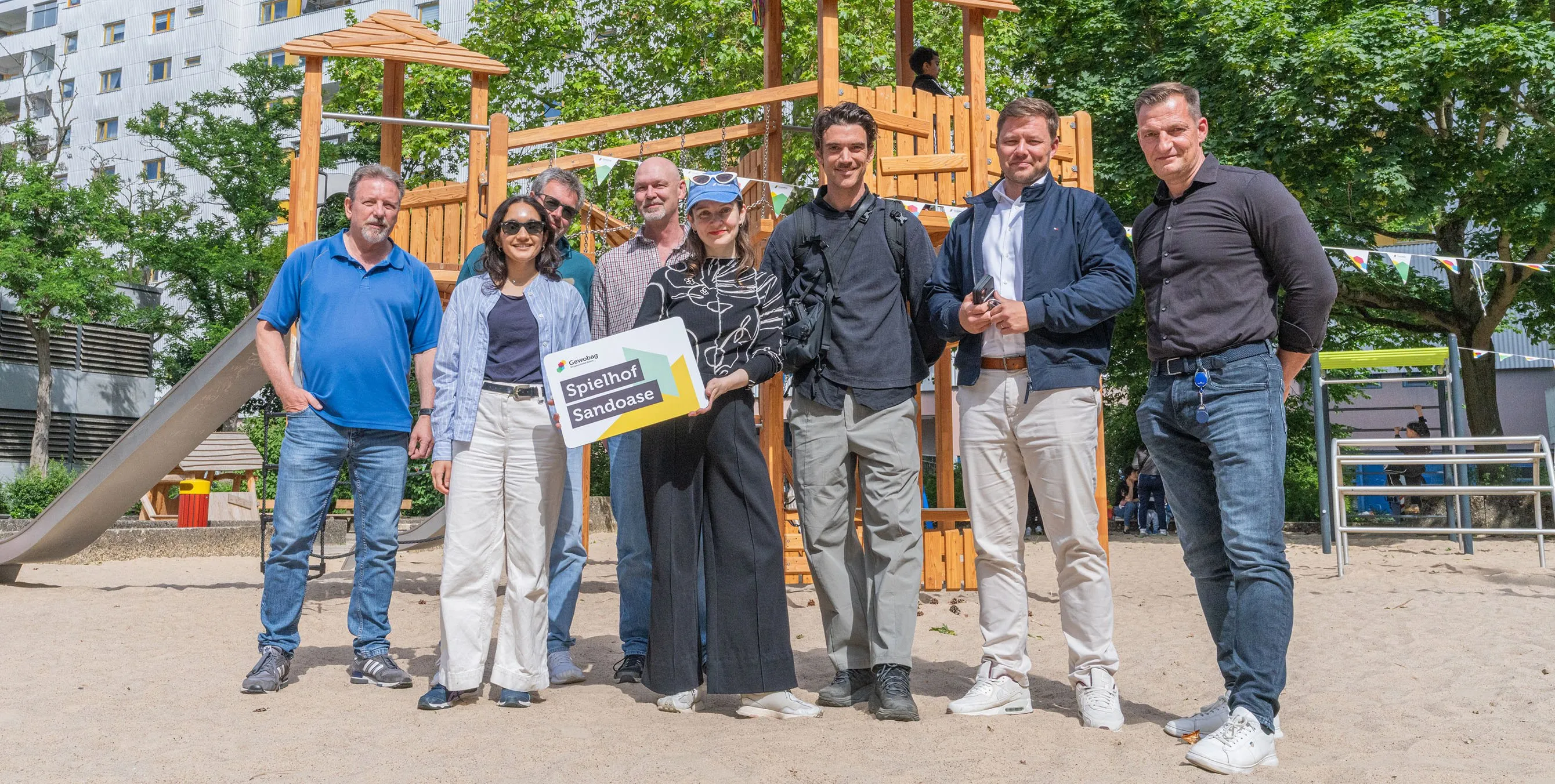 Acht erwachsene Personen stehen auf einem Spielplatz und lächeln in die Kamera. Eine mittig platzierte Frau hält ein Schild mit der Aufschrift "Spielhof Sandoase" in die Kamera. Foto: Felix Seyfert