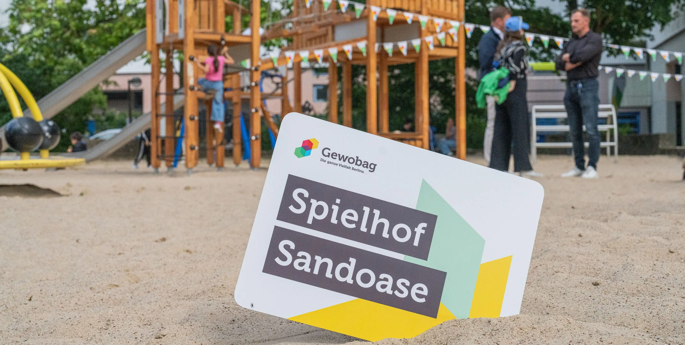 Im Hintergrund ist ein Spielplatz zu sehen, im Vordergrund ein Schild, das im Sand steckt. Aufschrift: Spielhof Sandoase