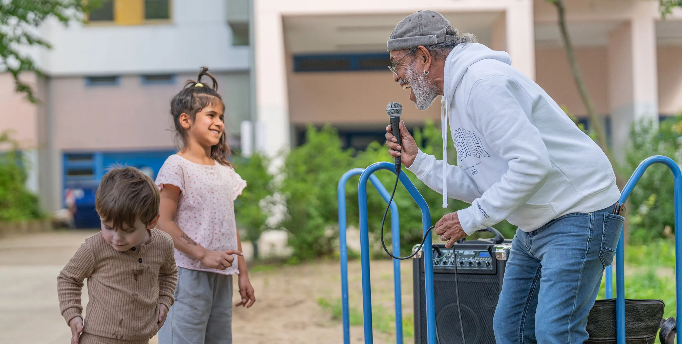 Ein älterer Musiker steht im Freien und singt lächelnd in ein Mikrofon. Zwei kleine Kinder stehen davor und schauen ihm zu. Foto: Felix Seyfert
