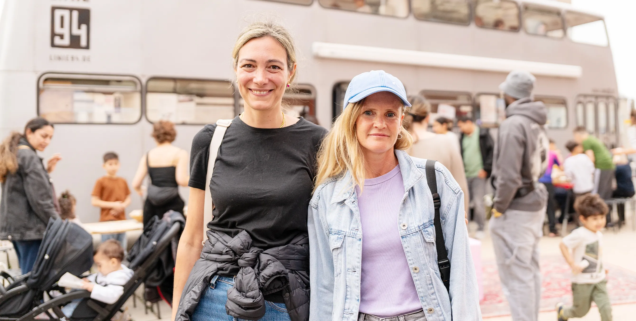 Zwei junge Frauen stehen vor einem Aktionsbus im Freien und lächeln in die Kamera. Im Hintergrund sind diverse Personen zu sehen. Foto: Ralph Maak