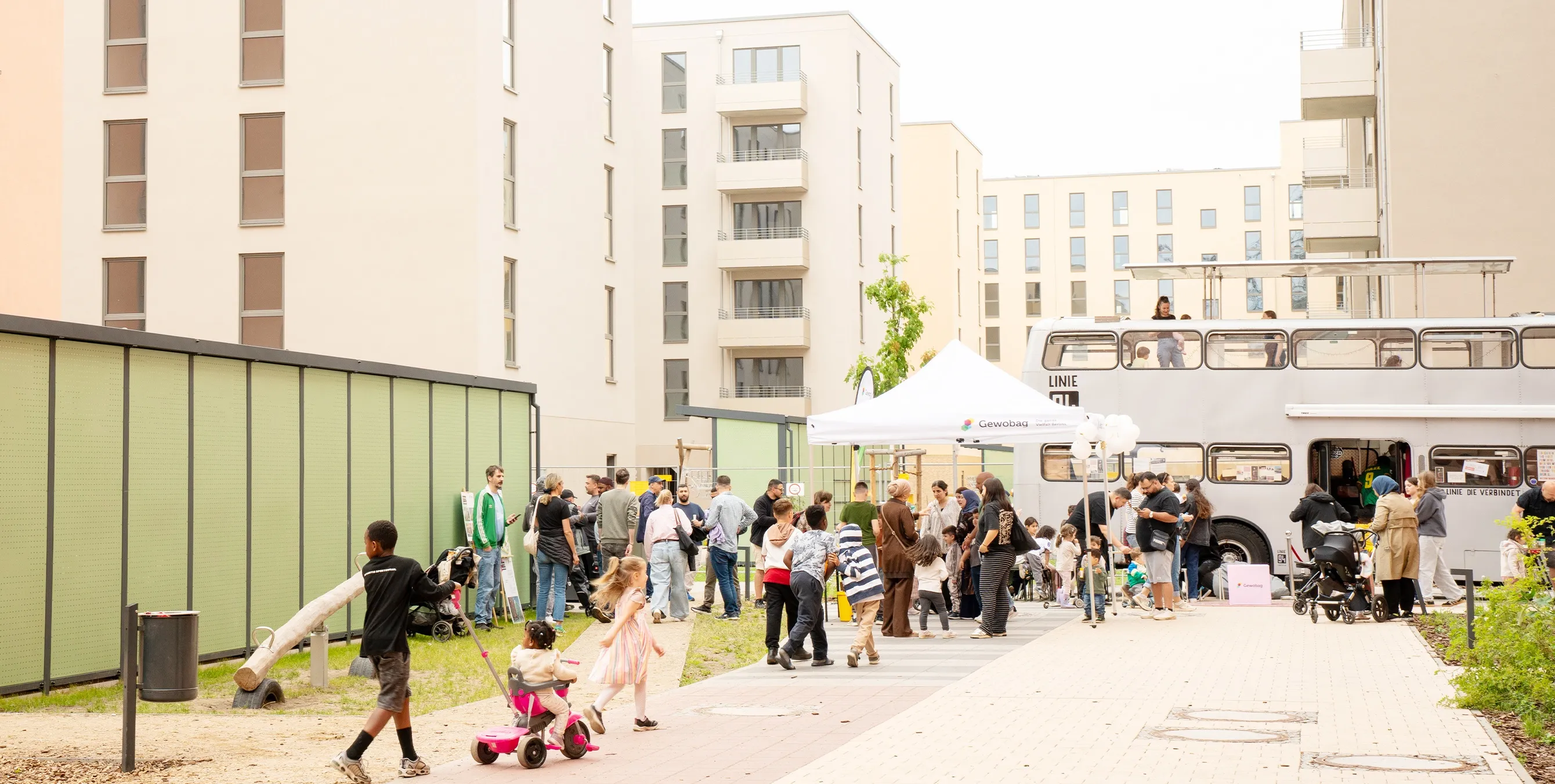 Aufnahme aus der Distanz: In einem Neubauquartier mit hellen Gebäuden findet ein Nachbarschaftsfest statt. Neben zahlreichen Menschen sind ein Pavillon und ein Aktionsbus zu sehen. Foto: Ralph Maak