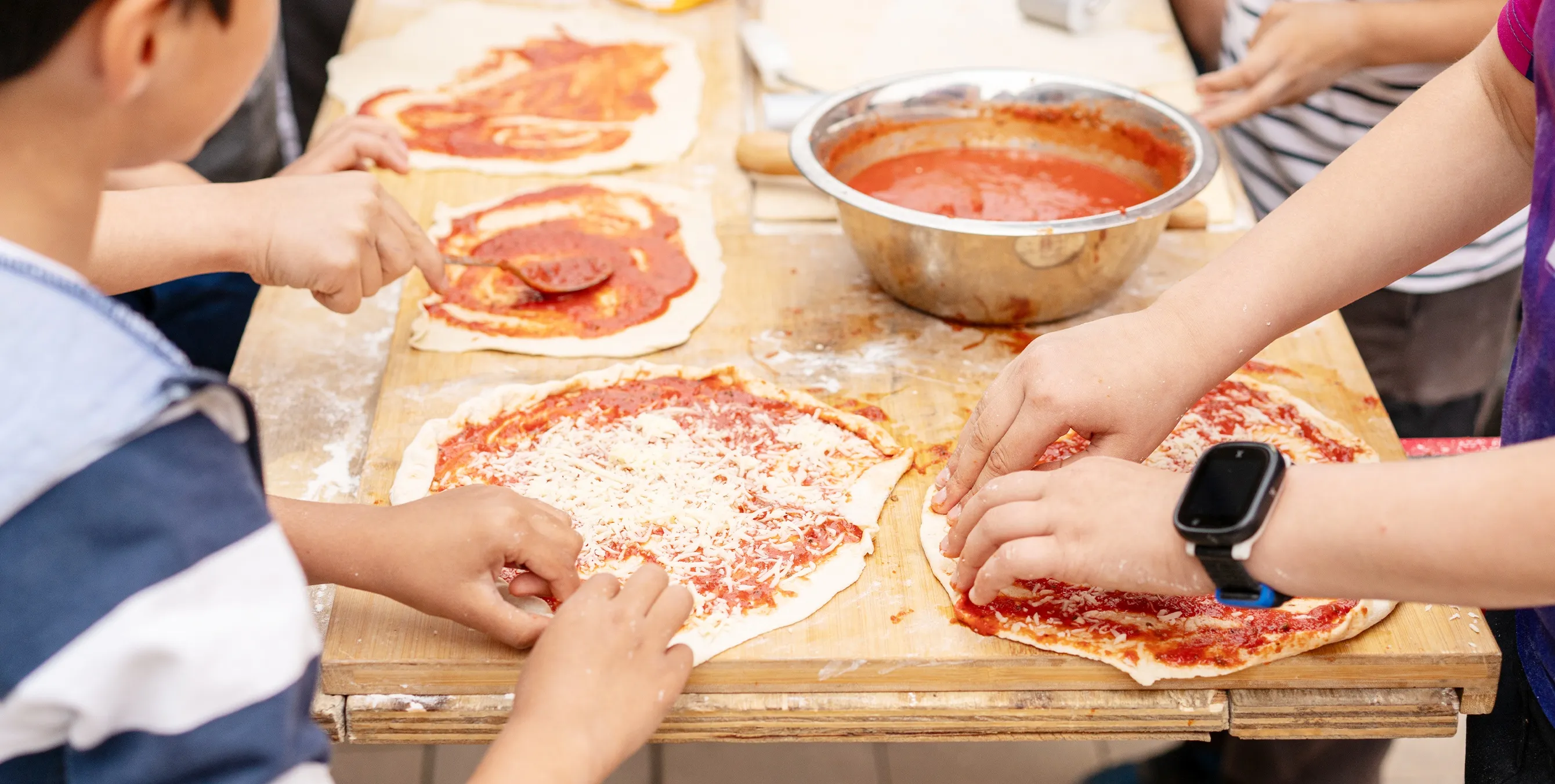 Großaufnahme: ein Tisch, auf dem von mehreren Kinderhänden Pizza zubereitet wird. Foto: Ralph Maak