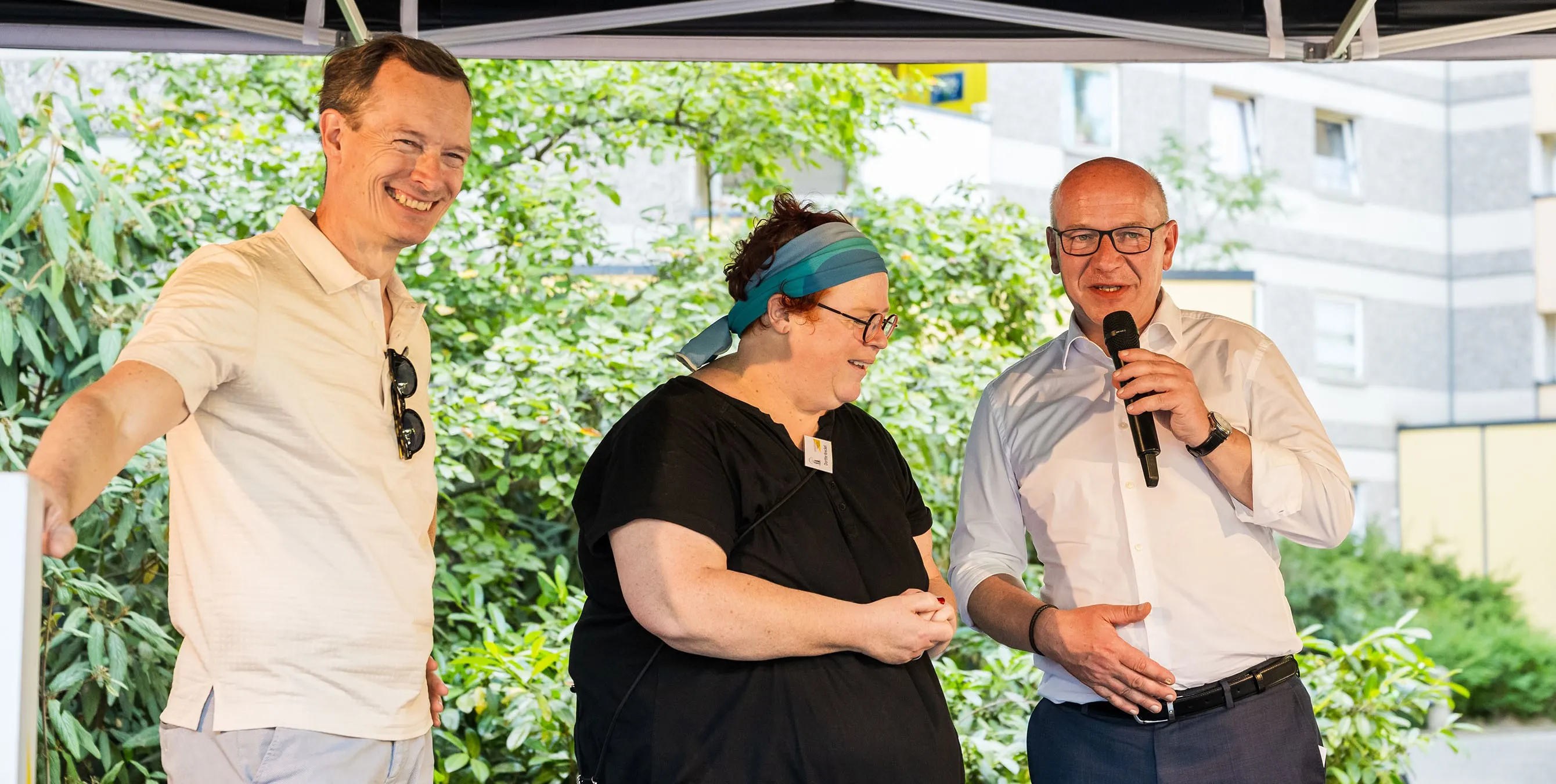 Zwei Männer und eine Frau auf einer kleinen Bühne bei einem Quartiersfest im Freien. Einer der Männer hält ein Mikrofon. Foto. City-Press GmbH
