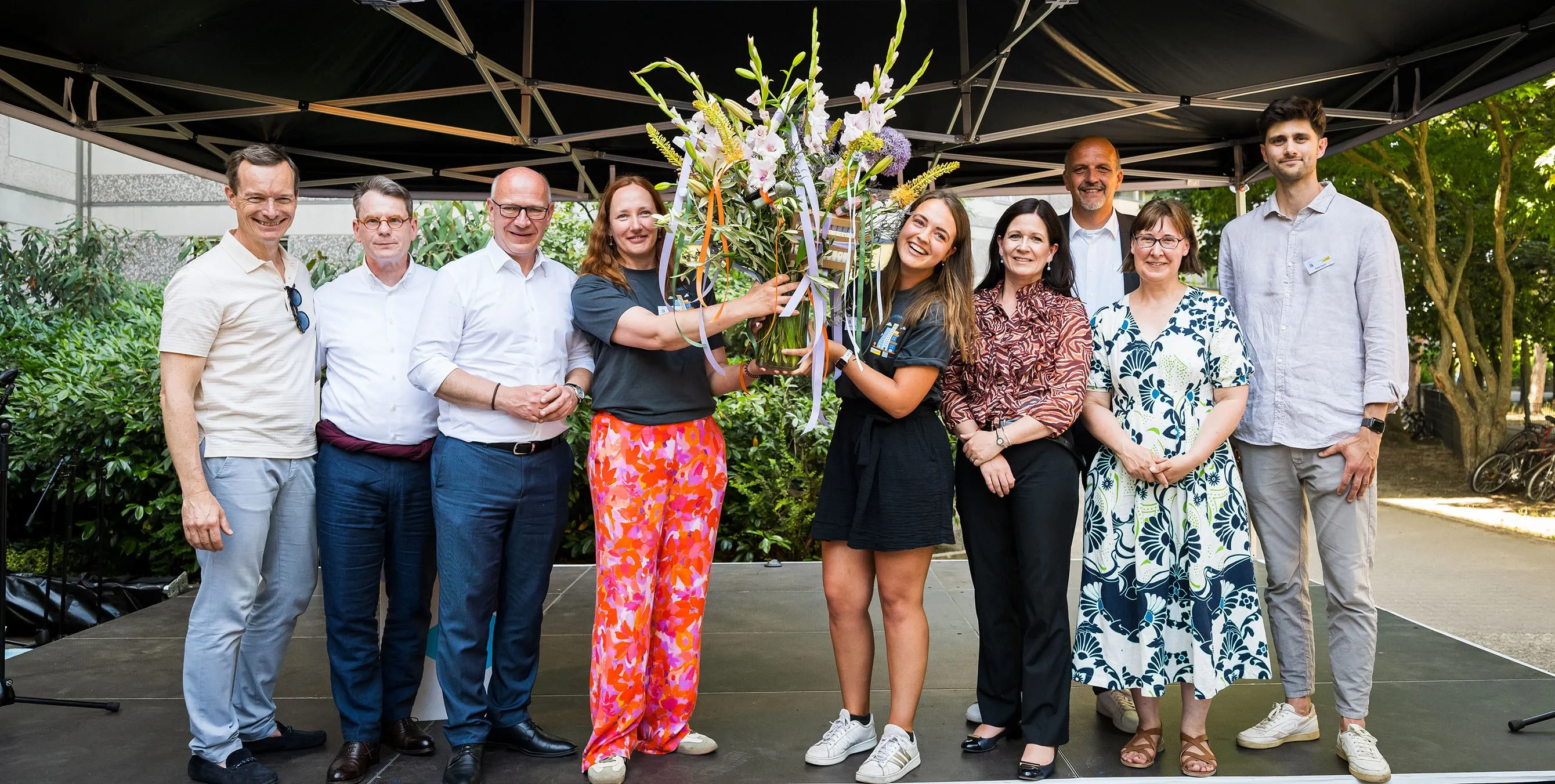 Zwie Frauen halten auf einer Bühne im Freien einen großen Blumenstrauß in der Hand und lächeln. Neben ihnen stehen sieben Personen. Gemeinsam posieren sie für ein Foto. Foto: City-Press GmbH