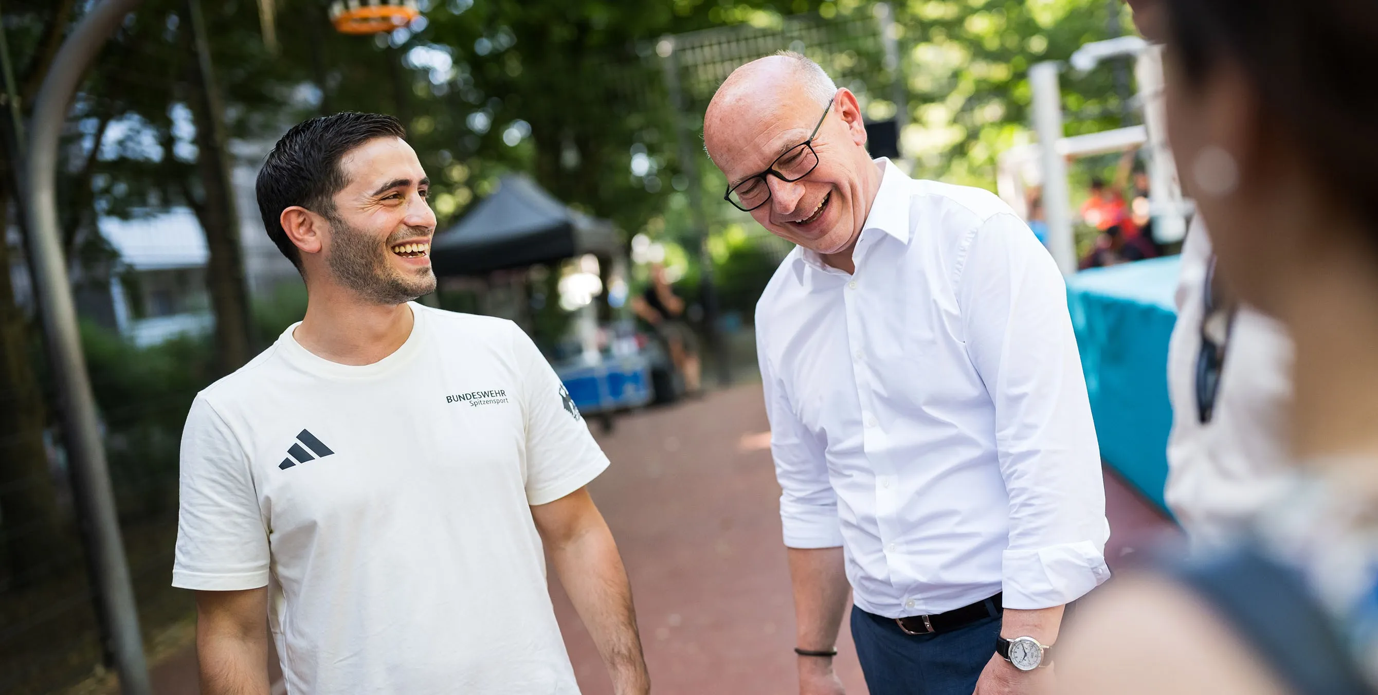 Ein jüngerer Sportler und ein älterer Mann stehen bei einem Quartiersfest im Freien und lachen miteinander. Foto: City-Press GmbH