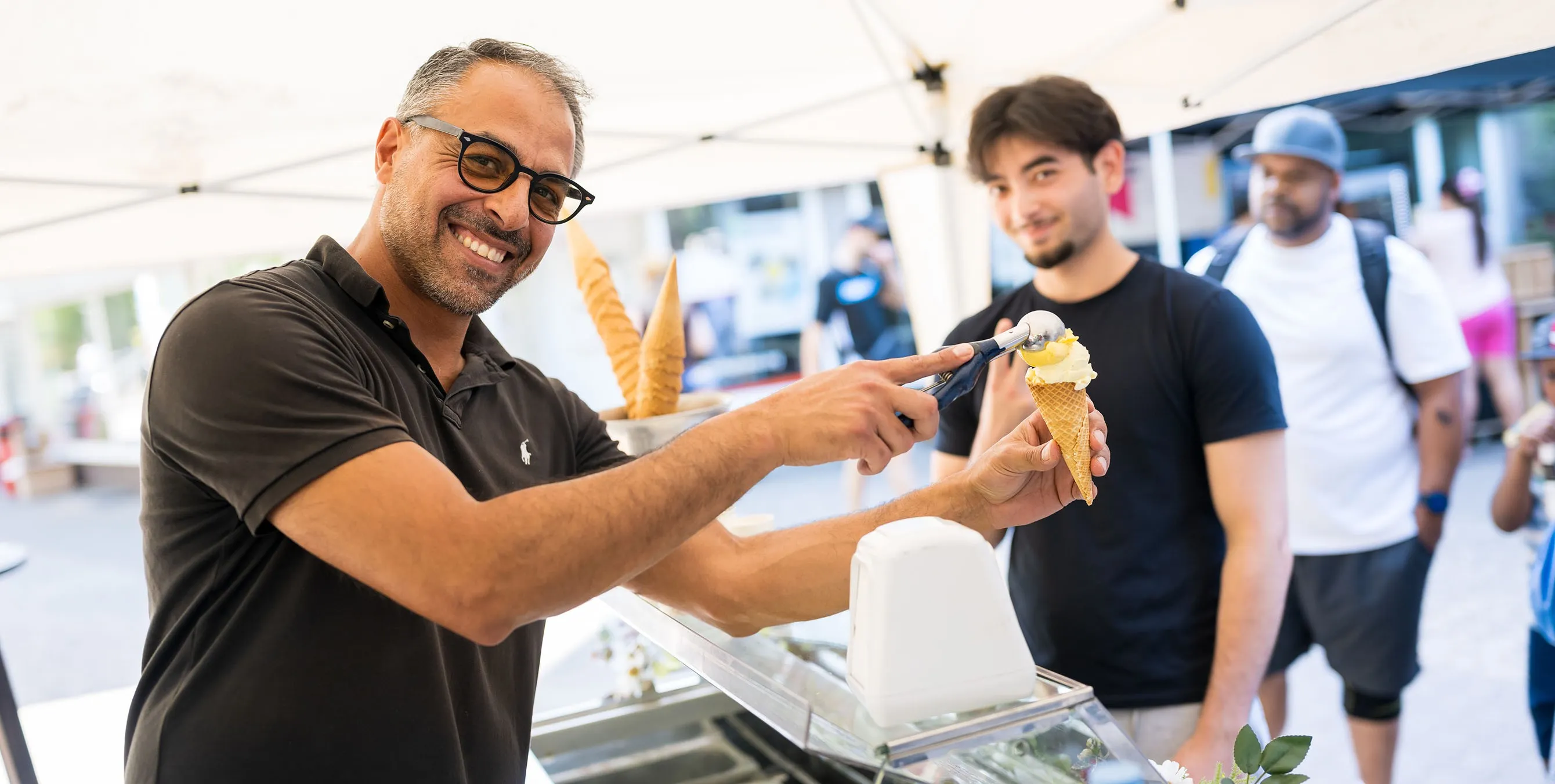 Szene von einem Quartiersfest: Ein Eisverkäufer setzt an seinem Eiswagen eine Eiskugel auf eine Waffel und lächelt in die Kamera. Foto: City-Press GmbH