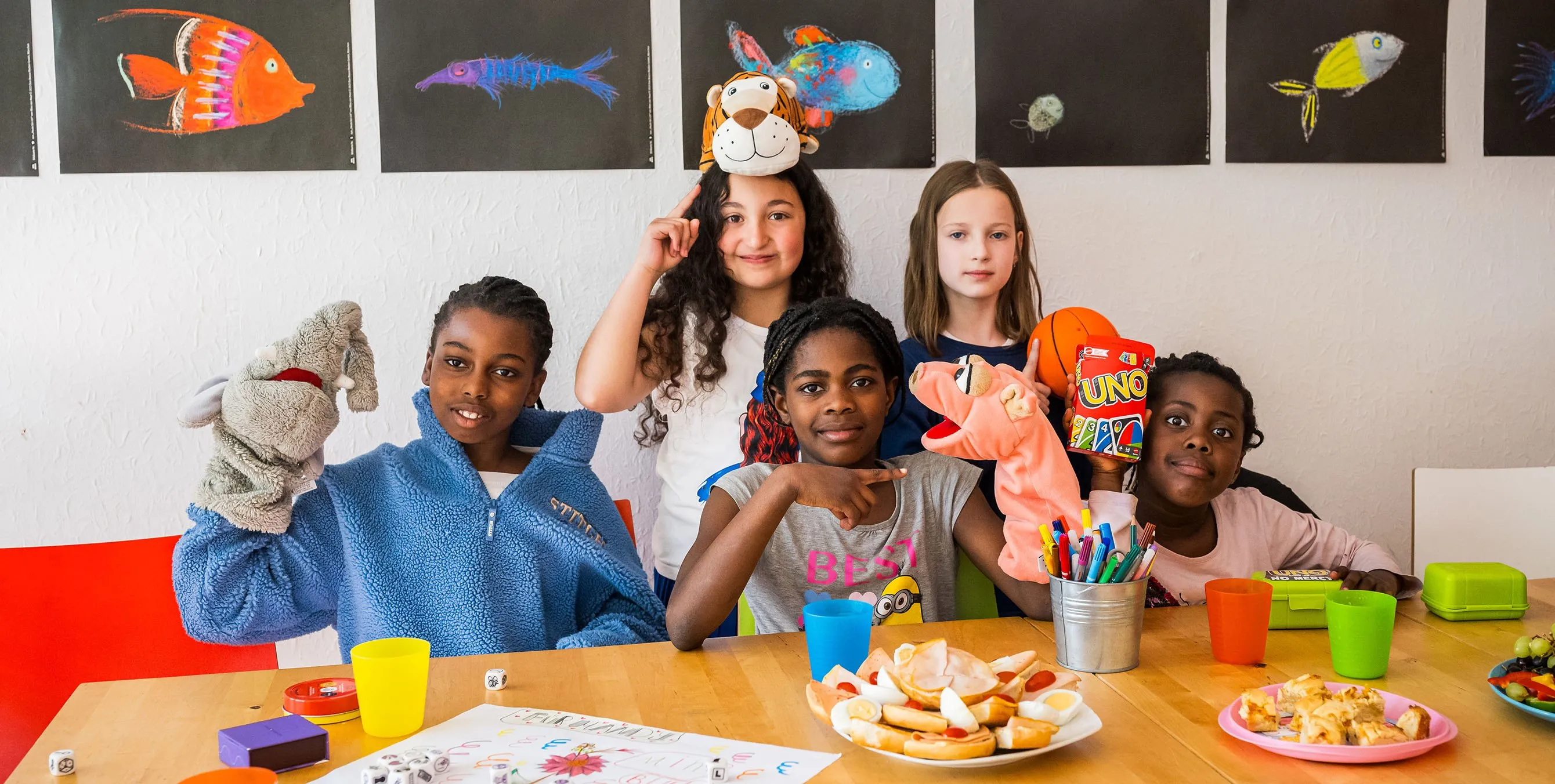 Fünf Mädchen sitzen in einer Wohnung an einem Tisch, auf dem Teller mit Essen, Trinkbecher, Spiele und Teller mit belegten Brötchen stehen. Die Mädchen tragen Handpuppen und schauen in die Kamera. Foto: City-Press GmbH