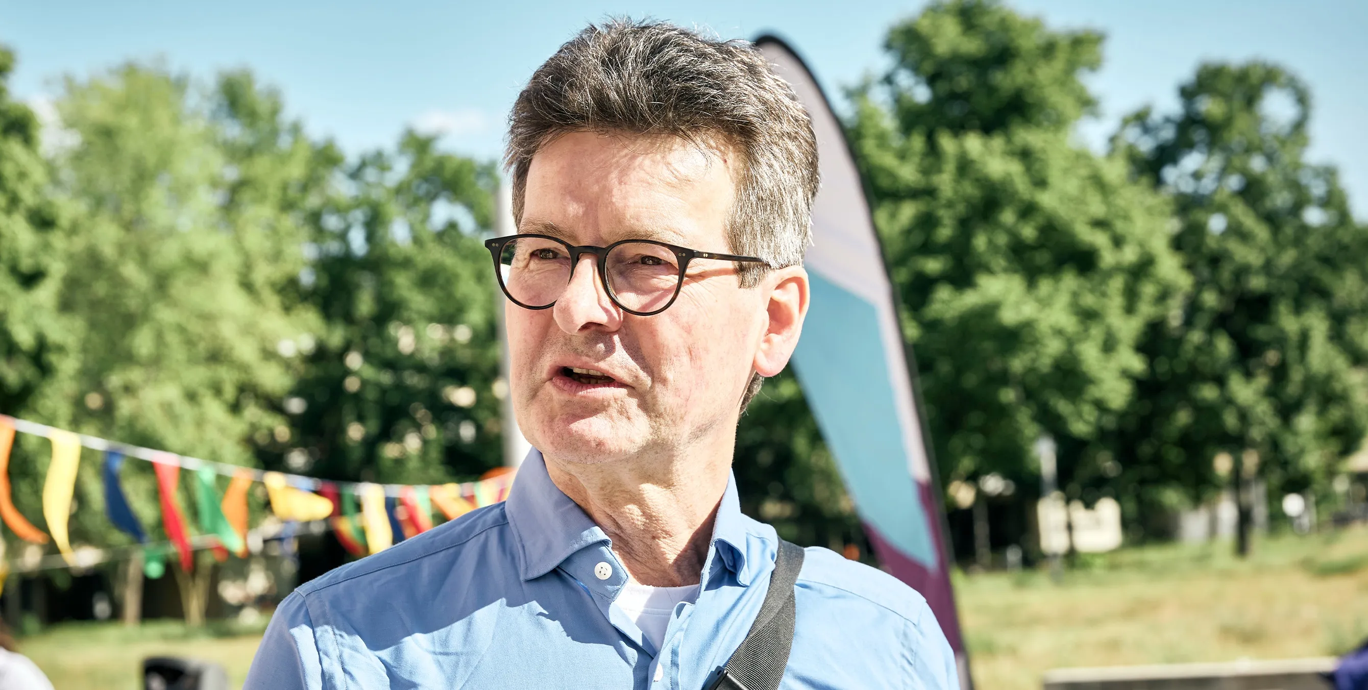 Ein Mann mit Brille steht im Freien und spricht. Im Hintergrund sind Wimpelketten, eine Beachflag, Bäume und eine Grünfläche zu sehen. Foto: Britta Leuermann