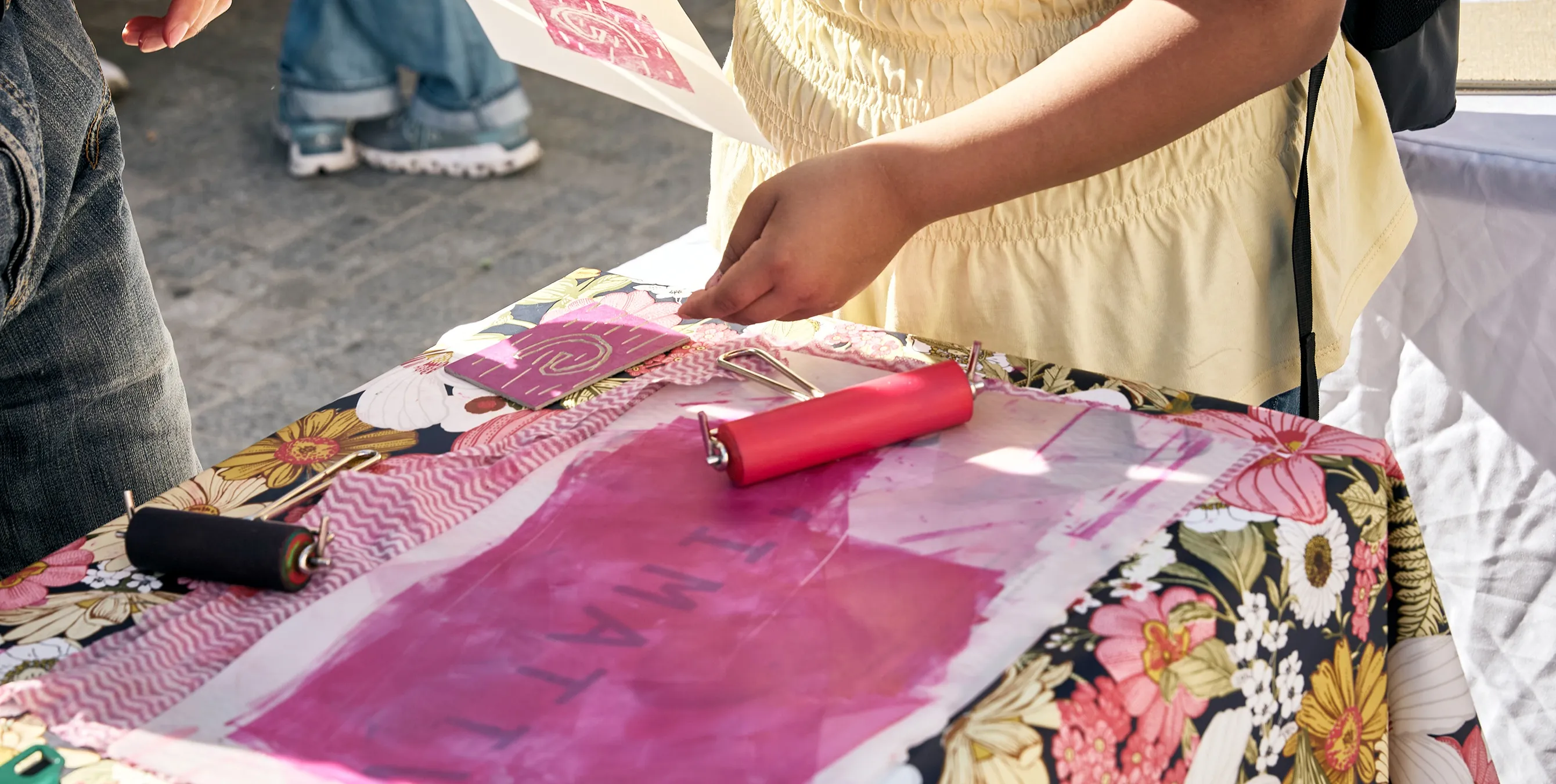Großaufnahme: Ein kleiner Tisch, auf dem bunten Papier und eine Rolle für Siebdruck liegen. Im Anschnitt ist ein Arm zu sehen, außerdem ein weißes Blatt mit einem farbigen Siebdruckmotiv. Foto: Britta Leuermann