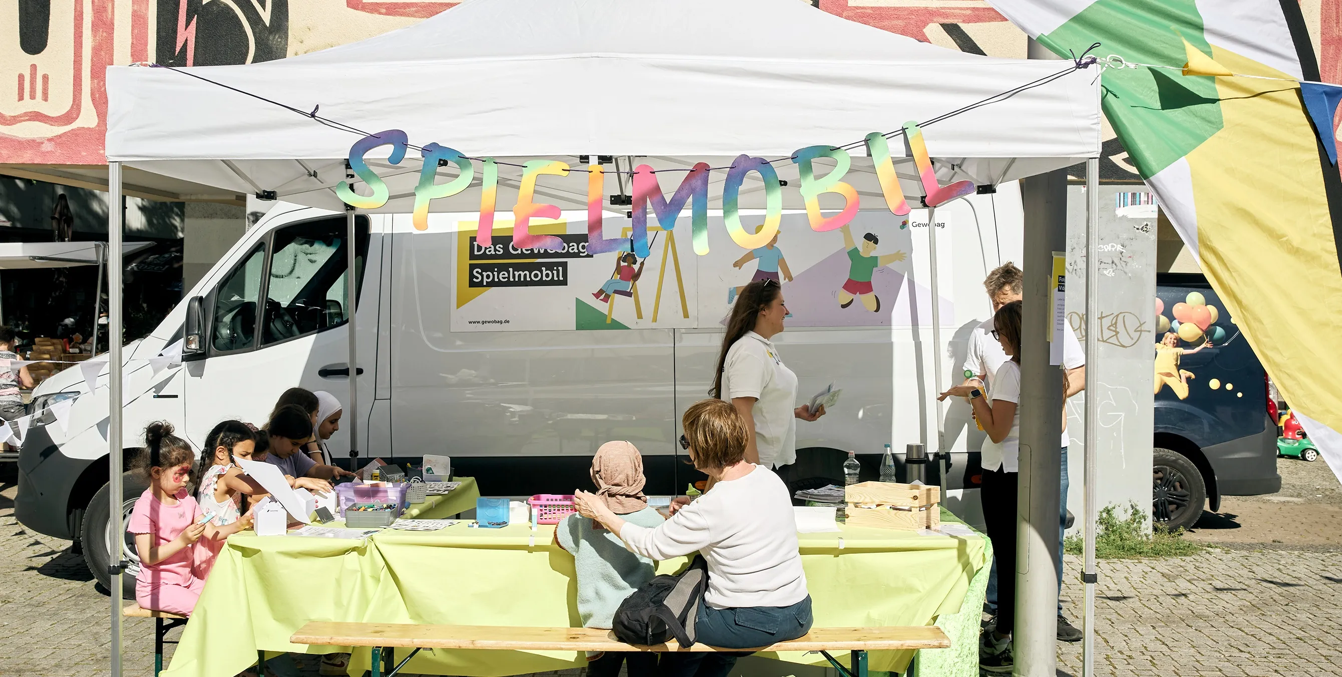 Kinder sitzen auf Bierbänken an einem Tisch mit Bastelutensilien, der unter einem Pavillon aufgebaut wurde. Im Hintergrund sieht man einen Van mit der Aufschrift "Das Gewobag Spielmobil", im Vordergrund eine Girlande mit dem Wort Spielmobil. Foto: Britta Leuermann