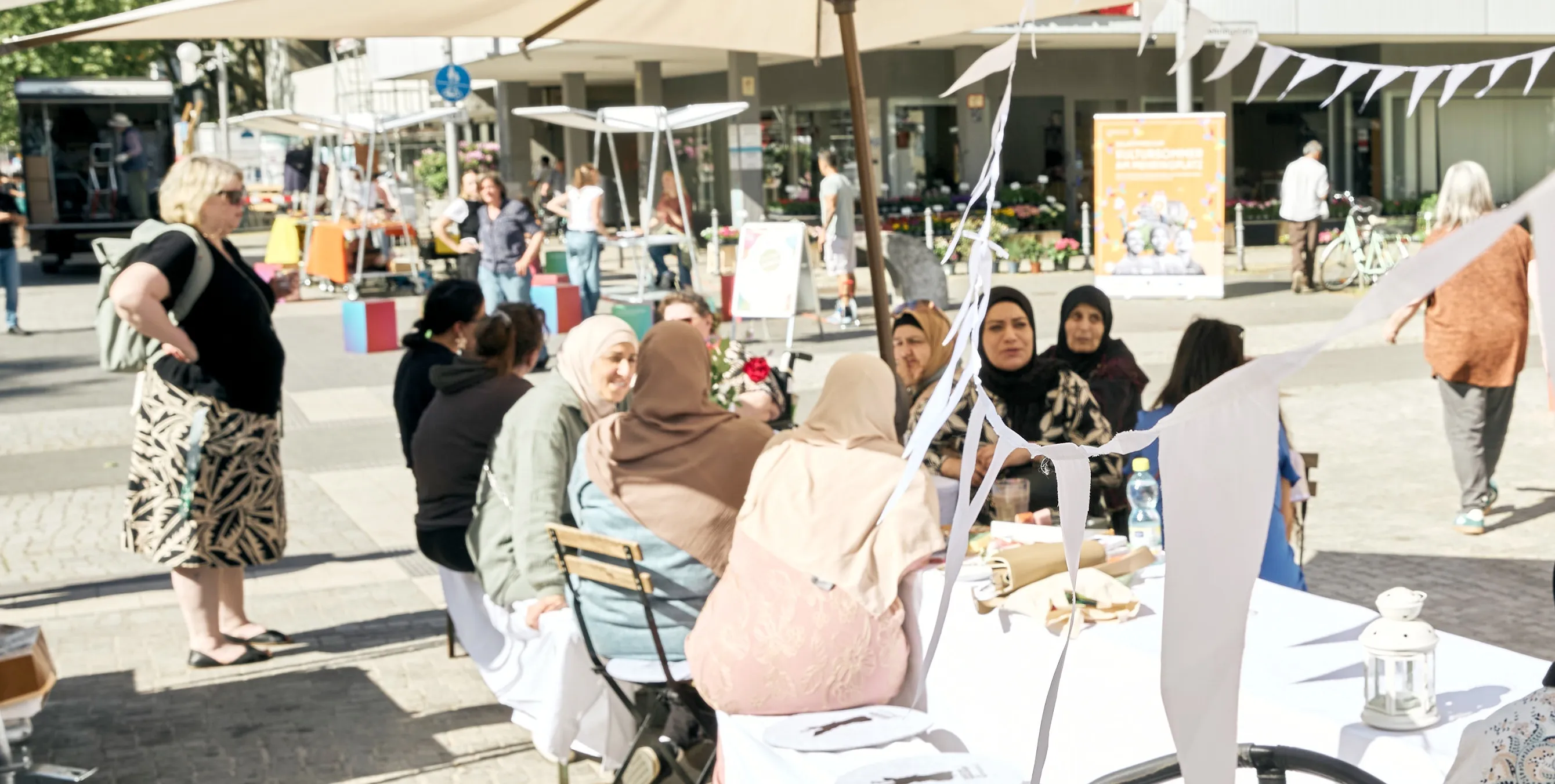 Szene auf einem öffentlichen Platz: Mehrere Frauen mit und ohne Kopftüchern sitzen an einer langen Tafel und unterhalten sich. Foto: Britta Leuermann
