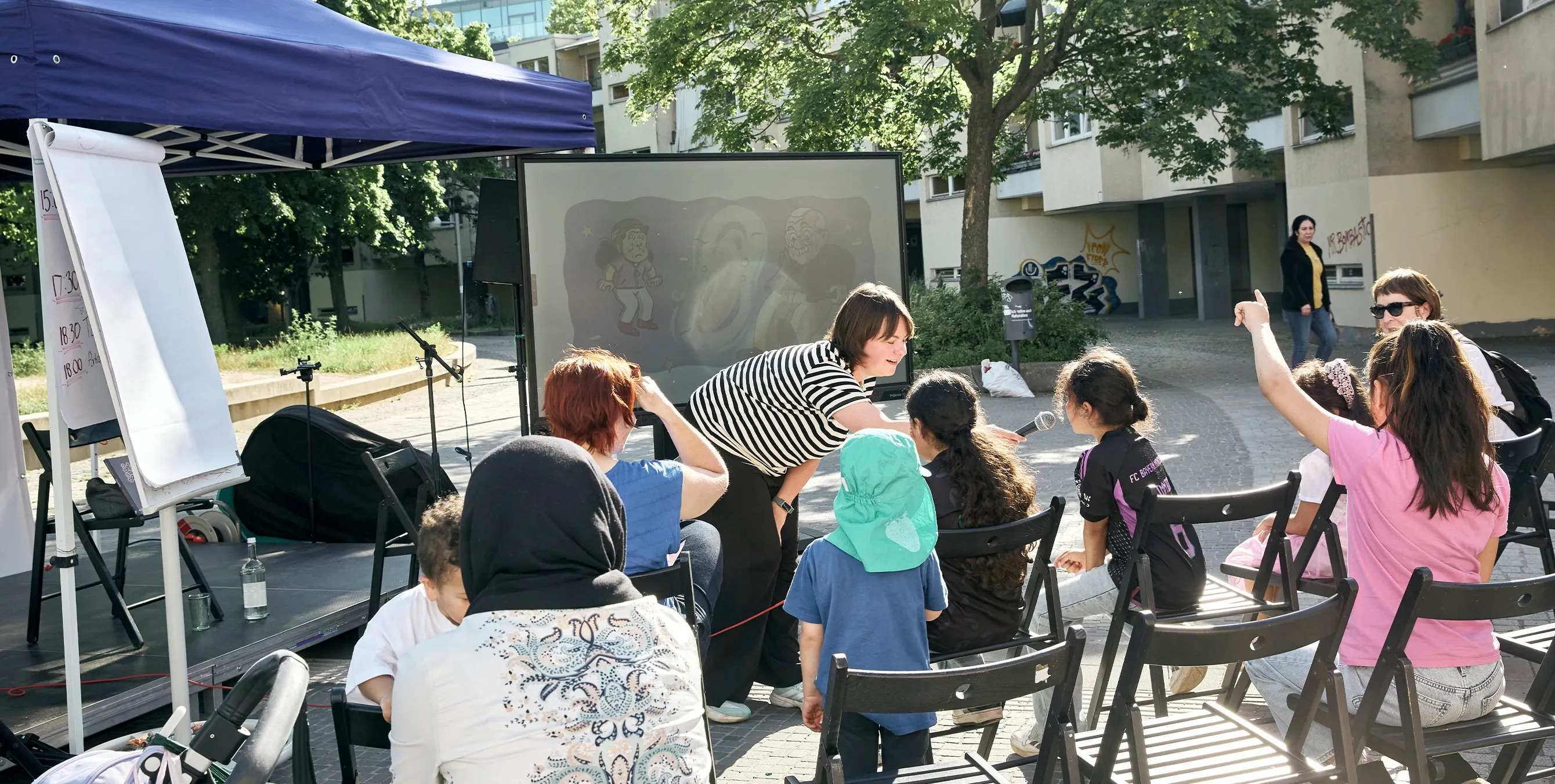 Szene auf einem öffentlichen Platz: Mehrre Kinder sitzen auf Stühlen vor einer Bühne. Eine Moderatorin mit Mikrofon stellt ihnen Fragen. Foto: Britta Leuermann