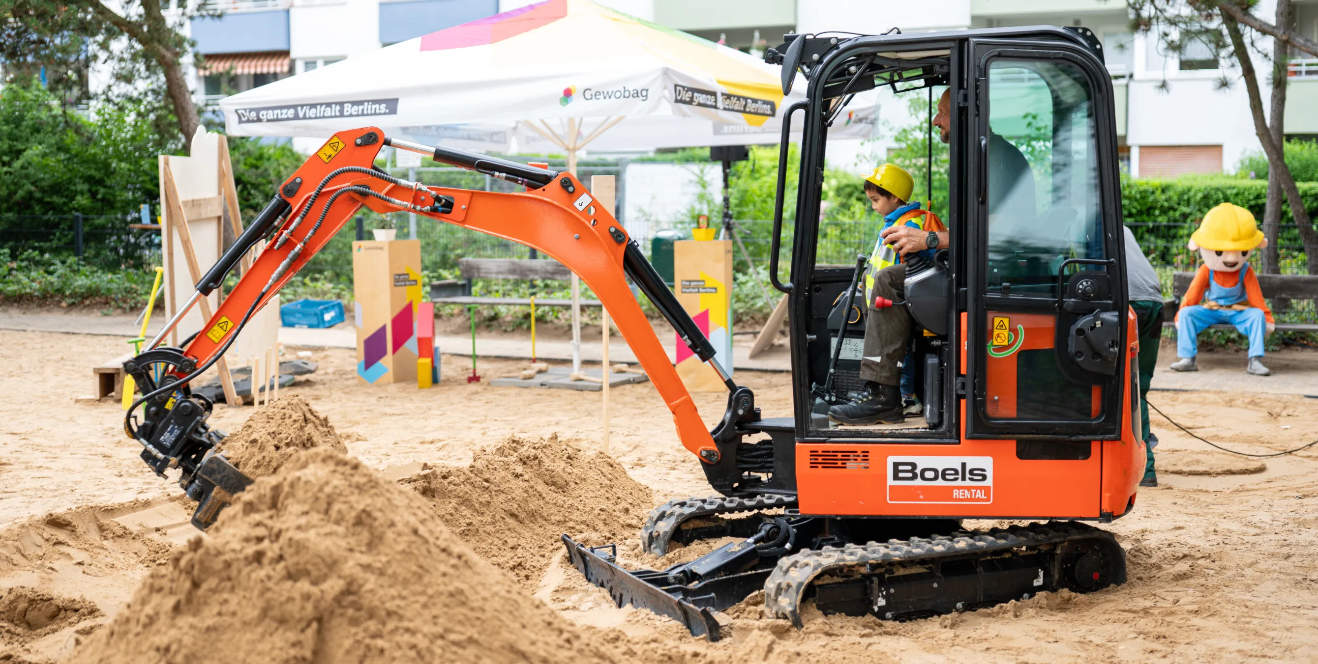Eine Fachkraft sitzt beim tag der kleinen Bauprofis mit einem Kind auf dem Schoß in einem Minibagger und schaufelt Sand. Foto: Ralph Maak