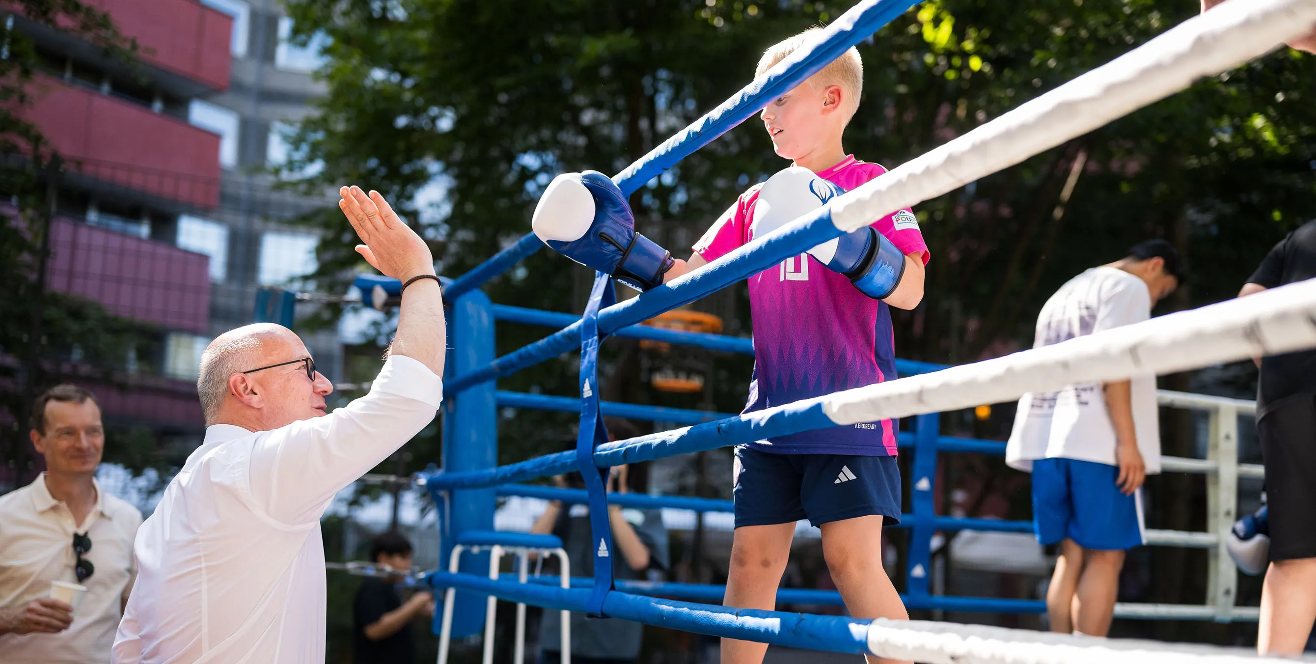 Ein Junge mit Boxhandschuhen steht in einem Boxring im Freien an den Seilen. Außerhalb steht ein Mann - beide klatschen durch die Ringseile miteinander ab. Foto: City-Press Gmbh