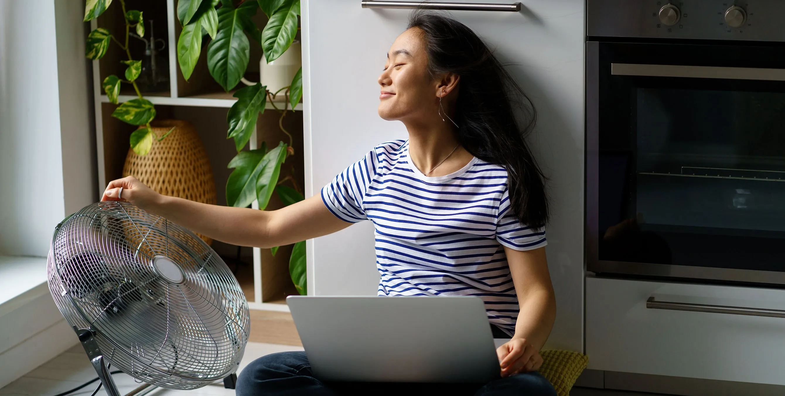 Eine junge Frau sitzt vor einer Küchenzeile auf dem Fußboden. Auf ihrem Schoß hat sie ein Notebook, neben ihr steht ein Ventilator. Foto: iStock