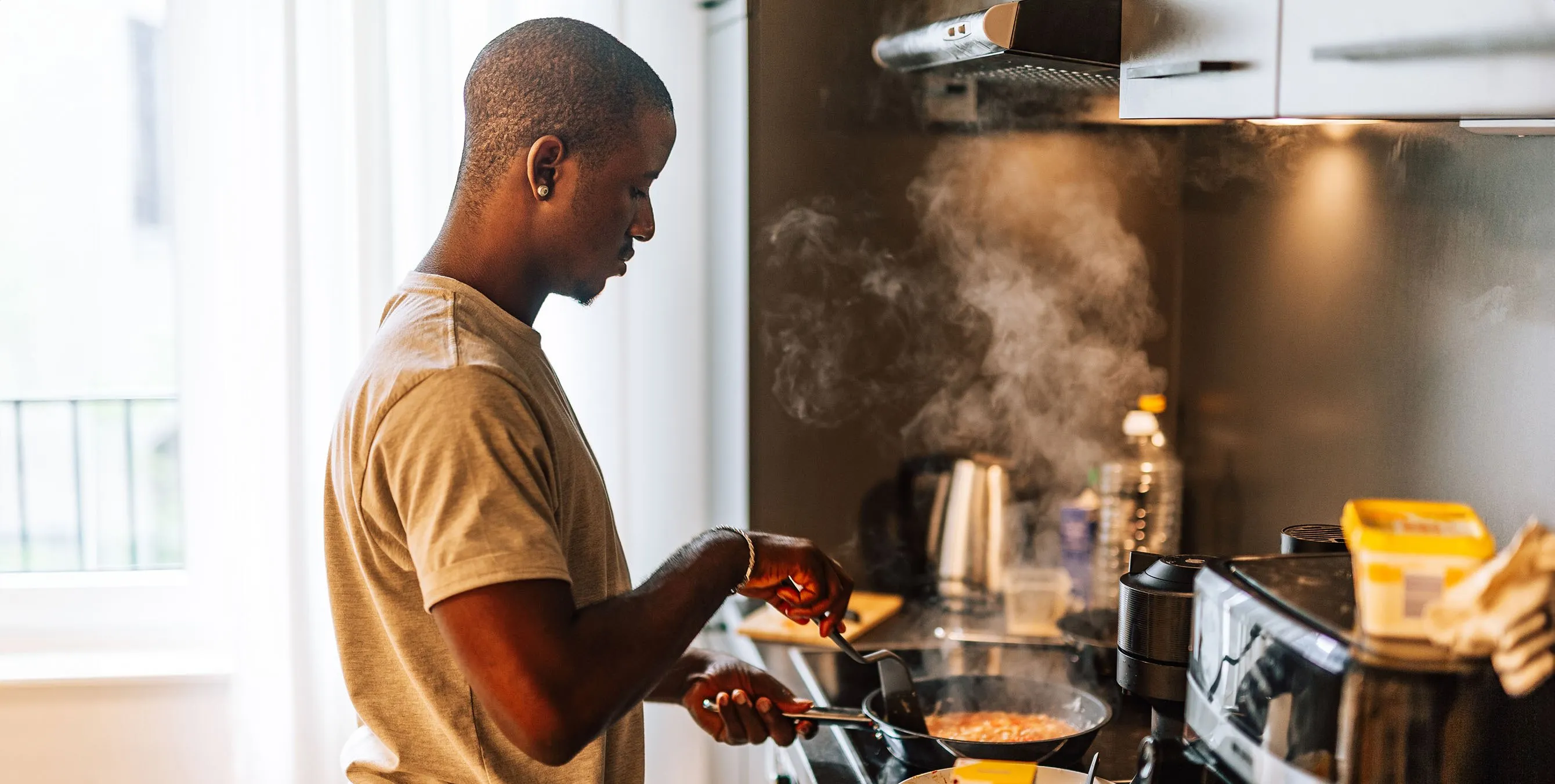 Ein junger Mann steht in einer Wohnung in der Küche und brät etwas in einer Pfanne. Es ist aufsteigender Dampf zu sehen. Foto: iStock