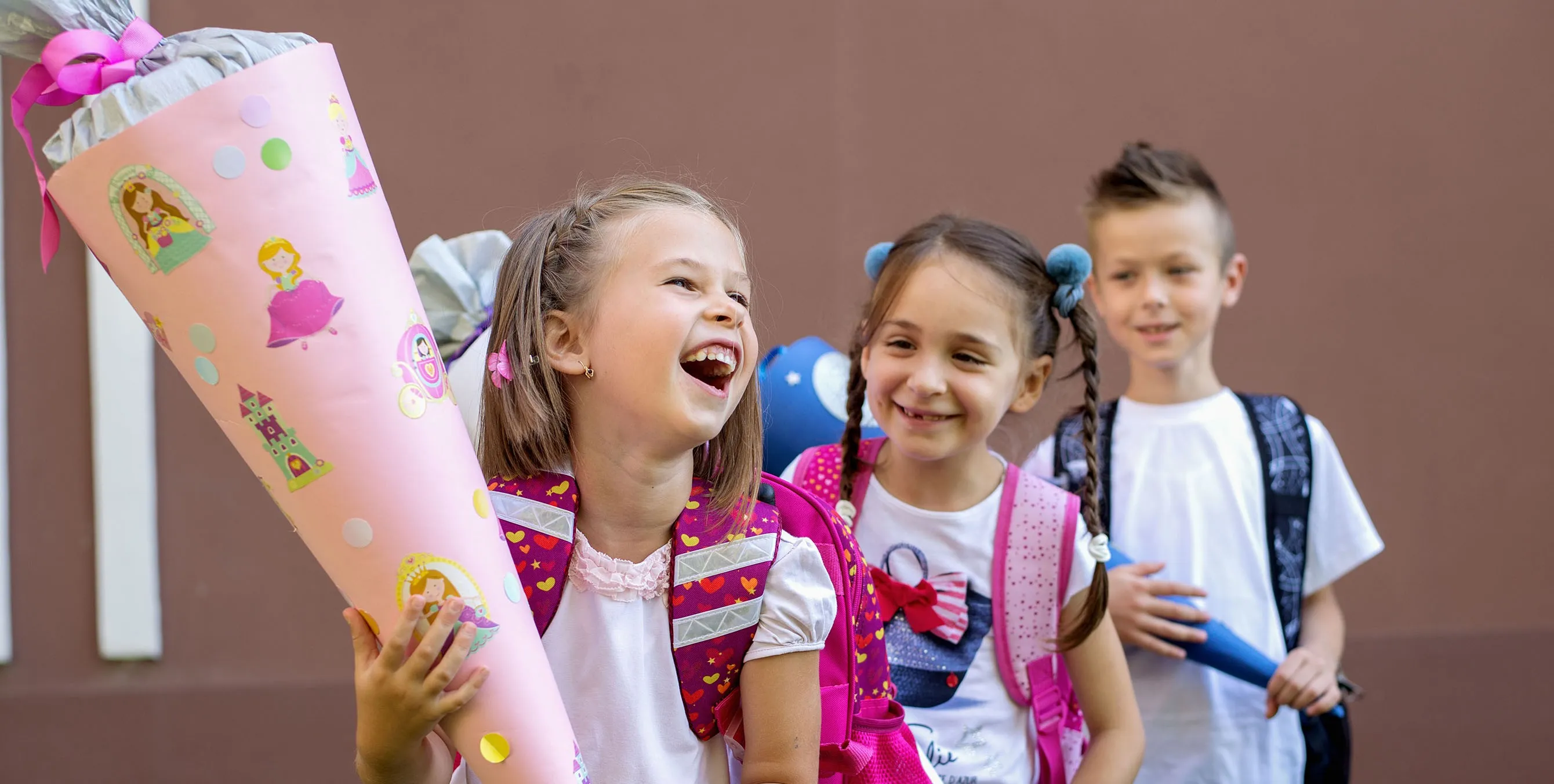 Zwei kleine Mädchen und ein kleiner Junge vor einer Wand. Alle halten Schultüten in der Hand und lachen oder lächeln. Foto: AdobeStock